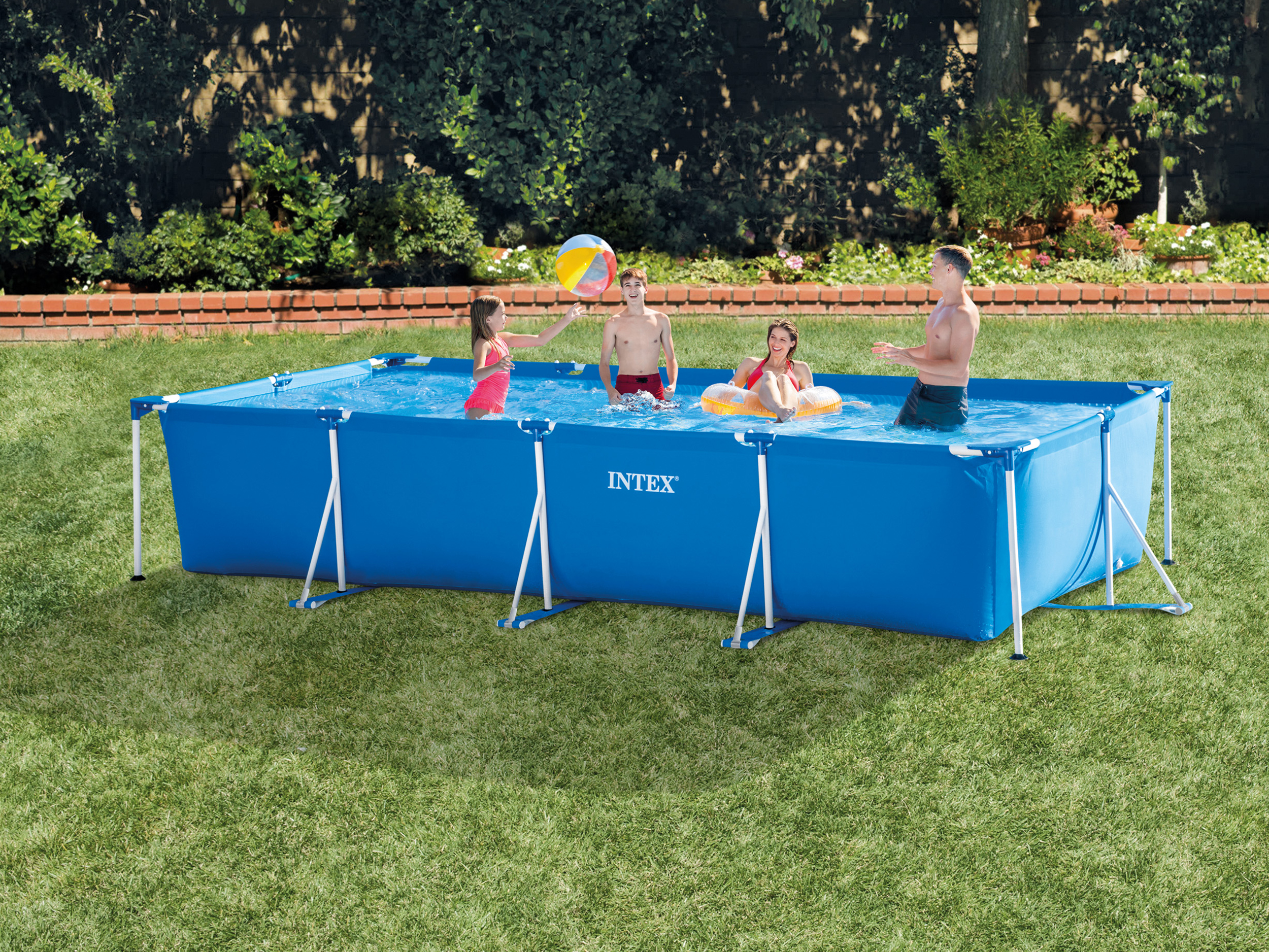Rodzina w niebieskim basenie Intex grająca piłką plażową. Zielona trawa, ceglany mur i krzaki w tle.
