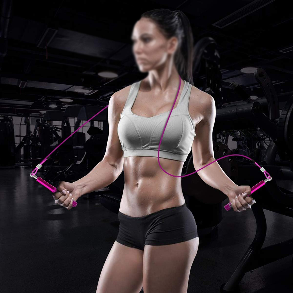 Eine Frau in Sportkleidung hält ein rosa Springseil in einem Fitnessstudio. Sie ist muskulös mit Bauchmuskeln und trägt schwarze Shorts und einen weißen Sport-BH.