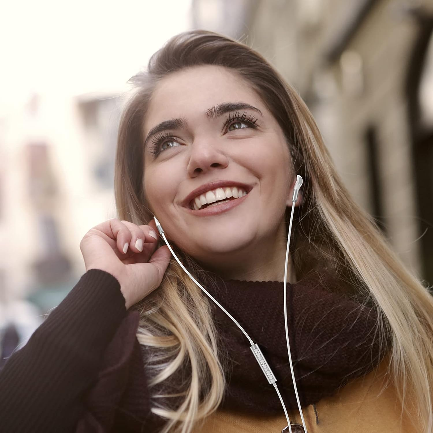 Blonde Frau lächelt, hört Musik mit weißen Kopfhörern. Sie trägt einen Schal und einen dunklen Mantel und blickt nach oben.