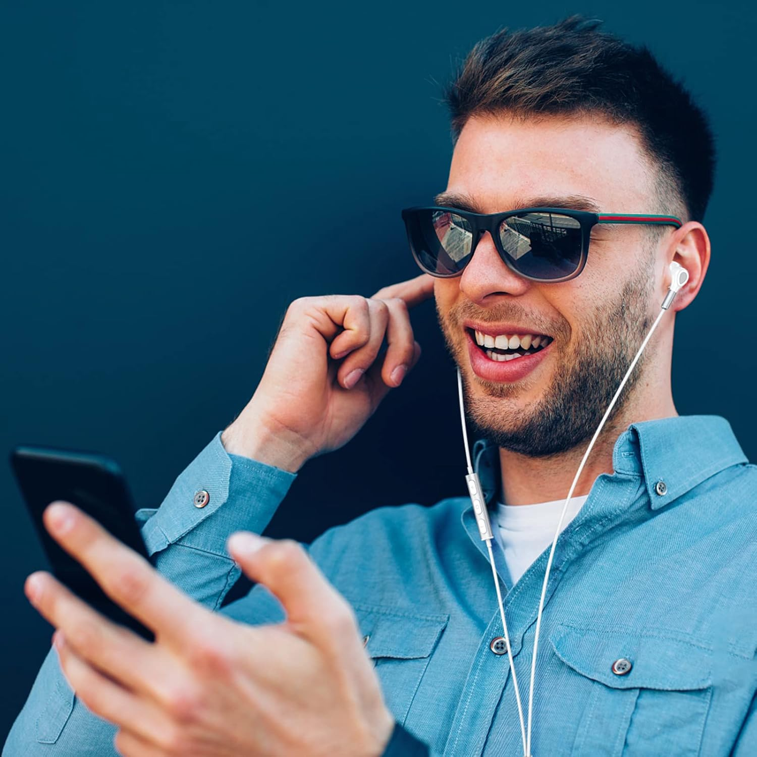 Ein Mann mit Sonnenbrille und Kopfhörern lächelt, während er ein Telefon hält. Er trägt ein blaues Hemd vor dunklem Hintergrund.