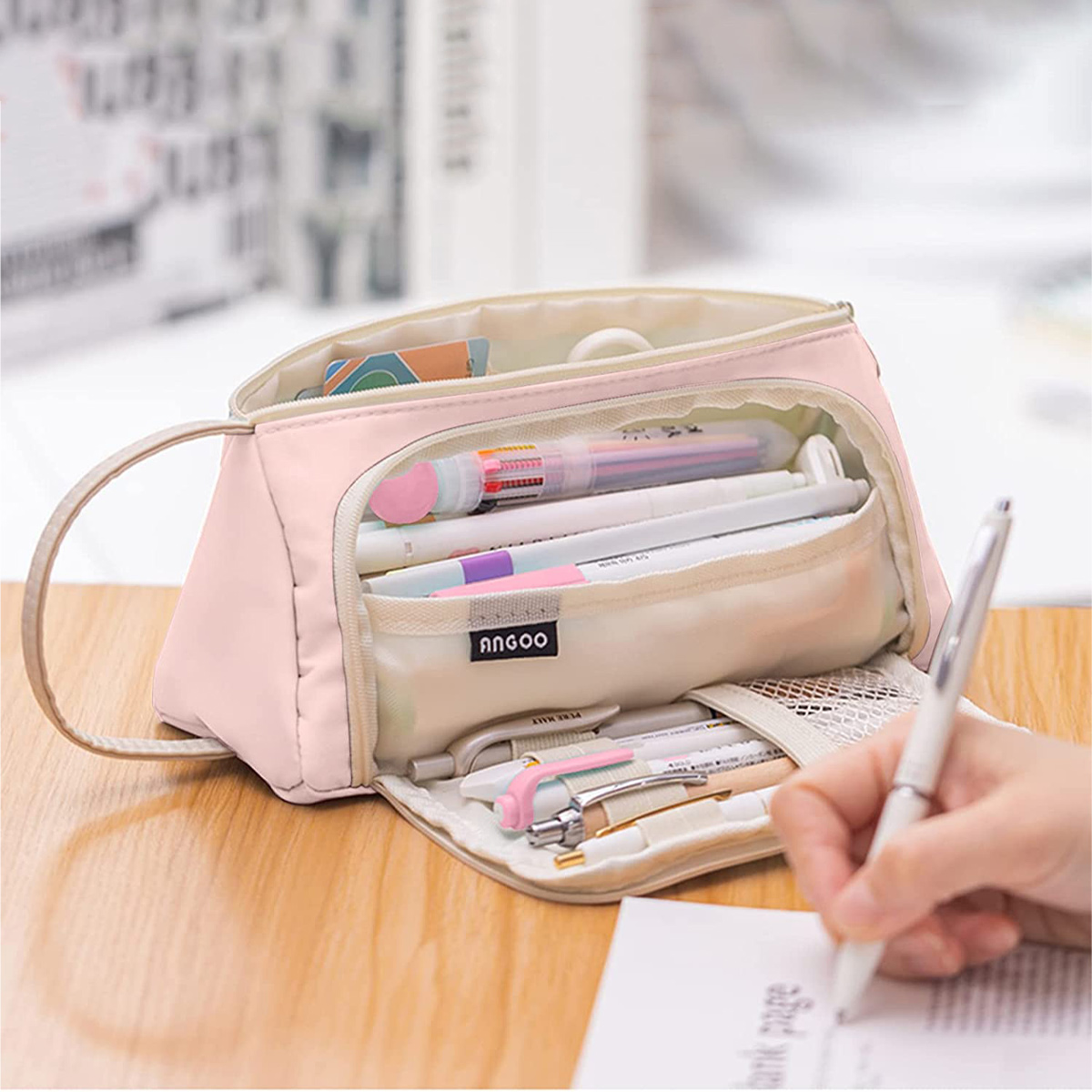 Ein rosa Federmäppchen ist geöffnet und mit Stiften und Schreibwaren gefüllt. Eine Hand schreibt mit einem Stift auf Papier.