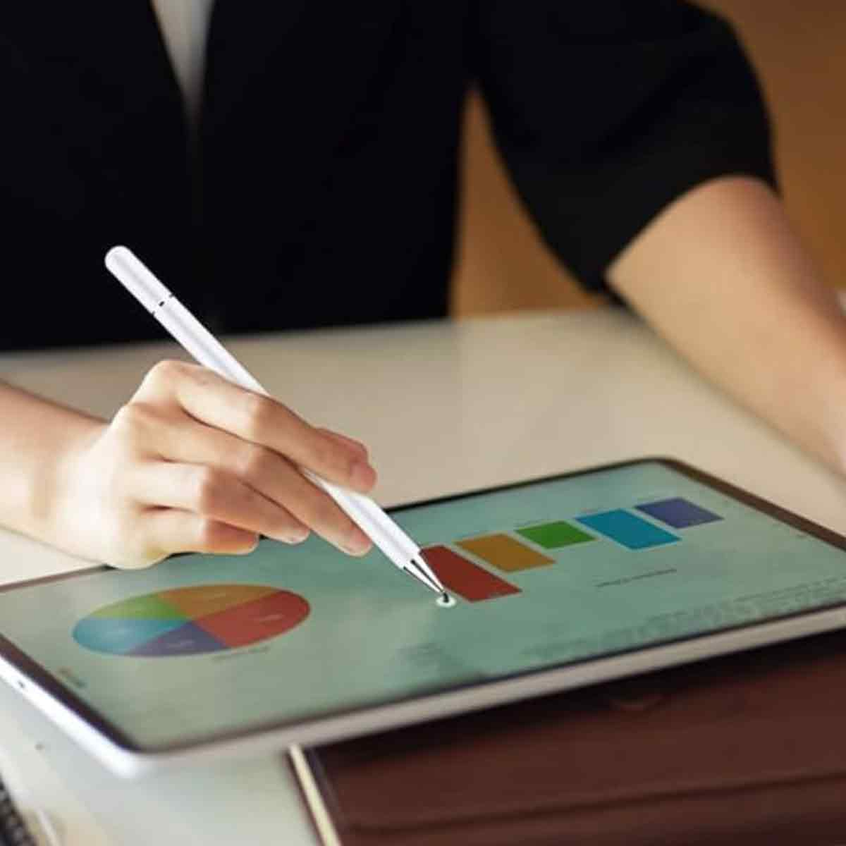 Eine Hand hält einen weißen Stift über einem Tablet, auf dem Diagramme angezeigt werden. Das Tablet liegt auf einem Tisch mit braunem Bezug.