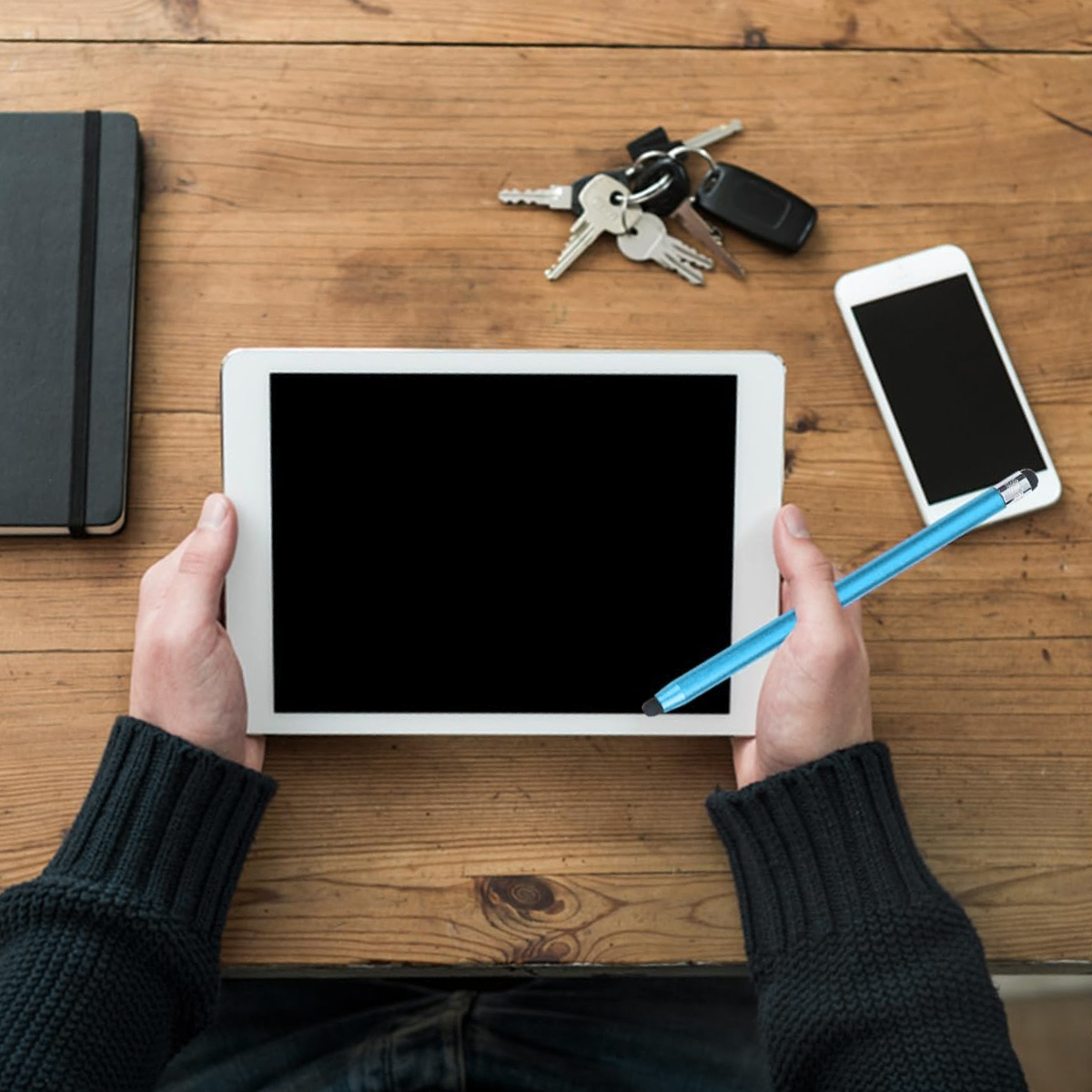 Hände halten ein Tablet mit schwarzem Bildschirm, einen blauen Stift, Schlüssel, ein Telefon und ein Notizbuch auf einem Holztisch.