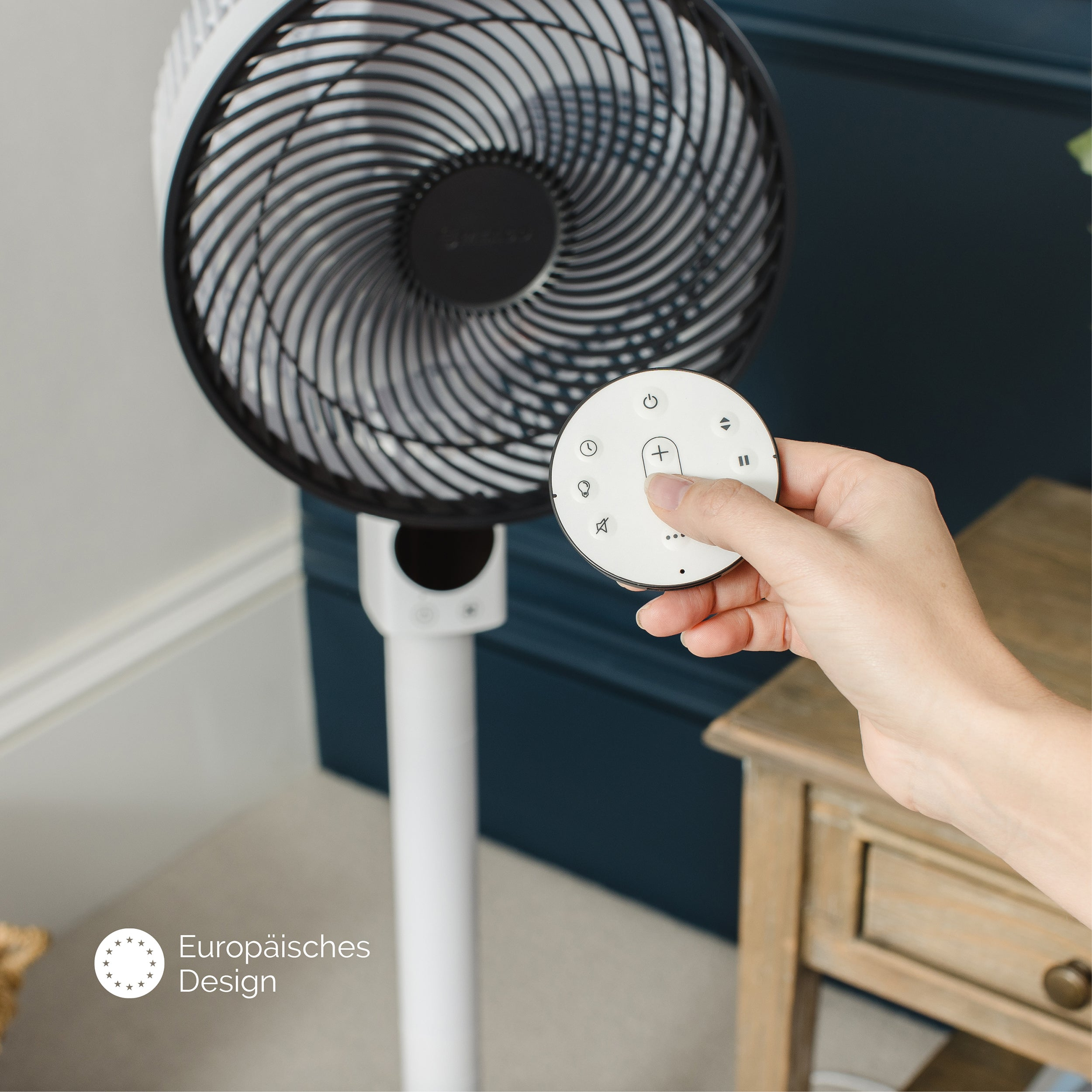 Ręka trzyma pilota do biało-czarnego wentylatora. Tło: drewniany stół i ciemnoniebieska ściana.