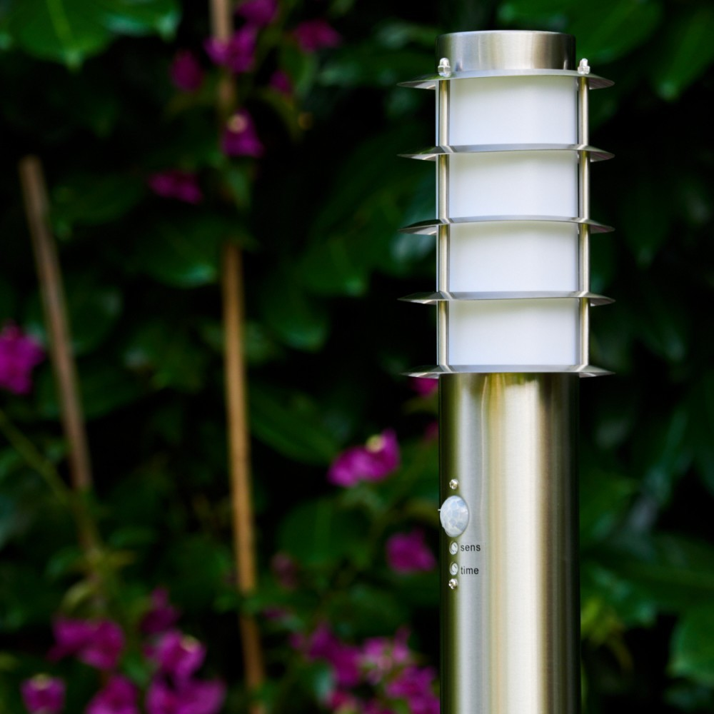 Eine metallene Außenlampe mit Sensor. Der Hintergrund hat grüne Blätter und lila Blumen.