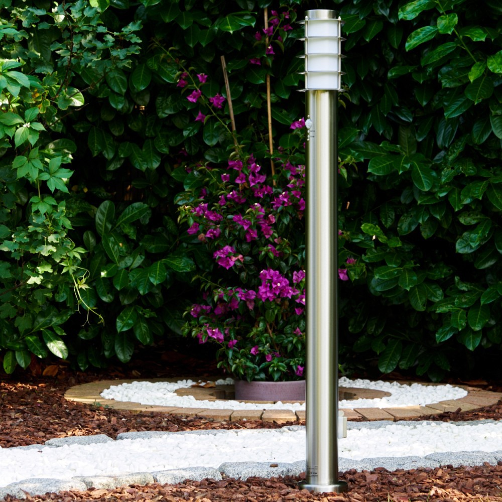 Eine Metalllaterne steht in einem Garten. Lila Blumen und grüne Büsche umgeben sie. Ein Steinweg führt zur Lampe.