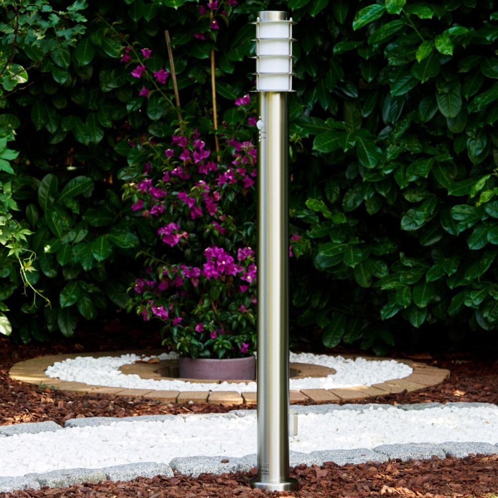 Eine Metall-Außenlampe steht in einem Garten. Hinter ihr stehen lila Blumen in einem Topf. Grüne Büsche bilden den Hintergrund.