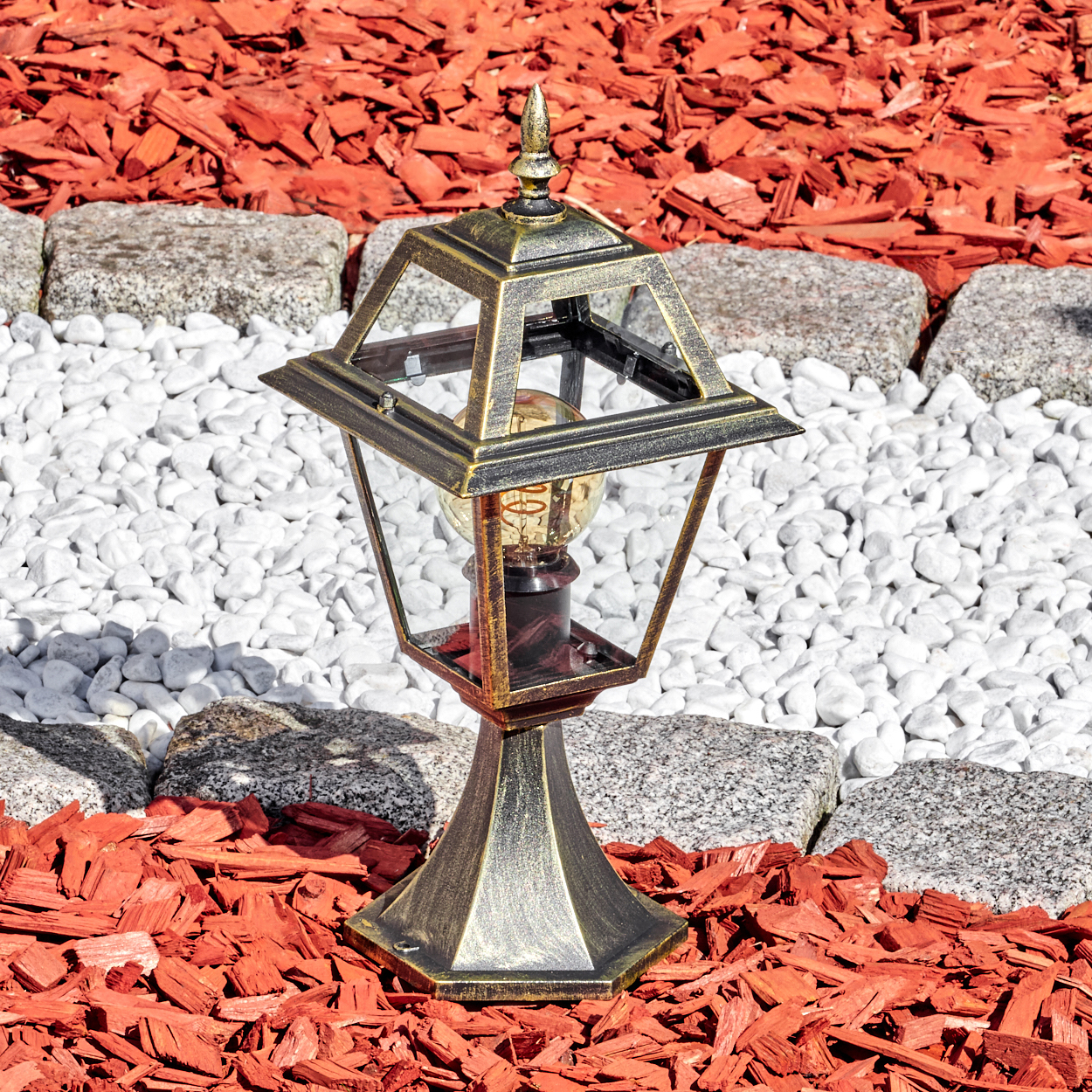 Eine bronzene Außenlampe mit einer Glühbirne im Inneren, die auf rotem Mulch, weißen Steinen und grauen Steinen steht.