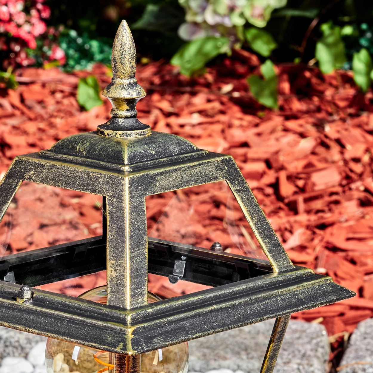 Eine dekorative Außenlaterne mit gold-schwarzem Finish. Der Hintergrund zeigt roten Mulch, Pflanzen und eine Steinoberfläche.