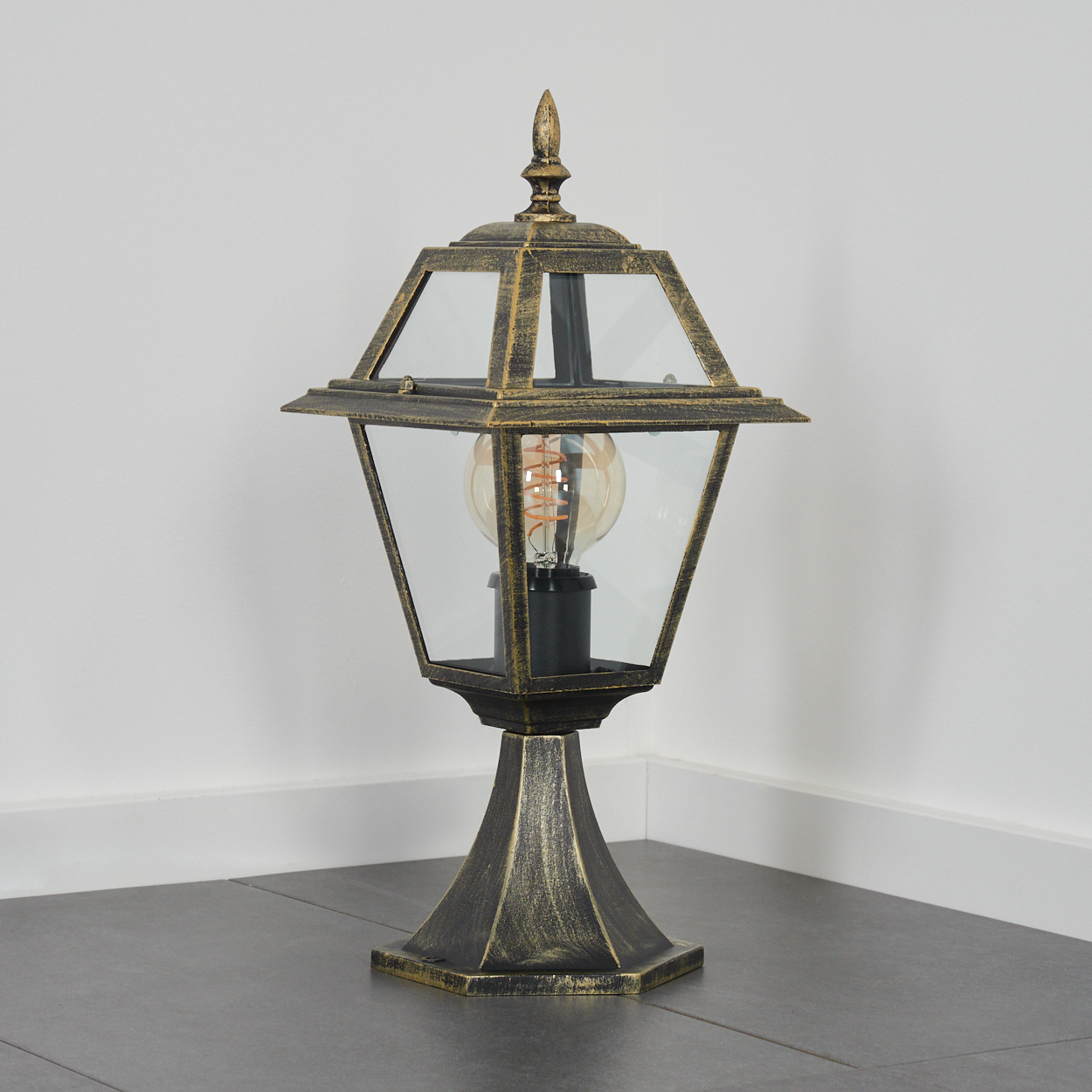 Eine verzierte bronzefarbene Außenlampe. Sie hat ein Glasgehäuse, eine leuchtende Glühbirne und steht auf einem sechseckigen Sockel.