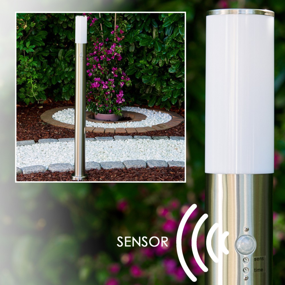 Eine moderne Außenlampe steht in einem Garten. Die Lampe hat einen zylindrischen weißen Aufsatz und einen Metallsockel. Der Hintergrund umfasst Pflanzen und einen Steinweg.