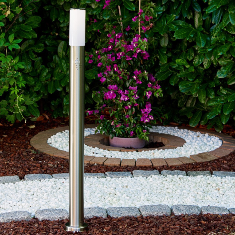 Eine Metall-Außenlampe steht in einem Garten. Rosa Blumen in einem Topf, weiße Kieselsteine und grüne Büsche umgeben die Lampe.