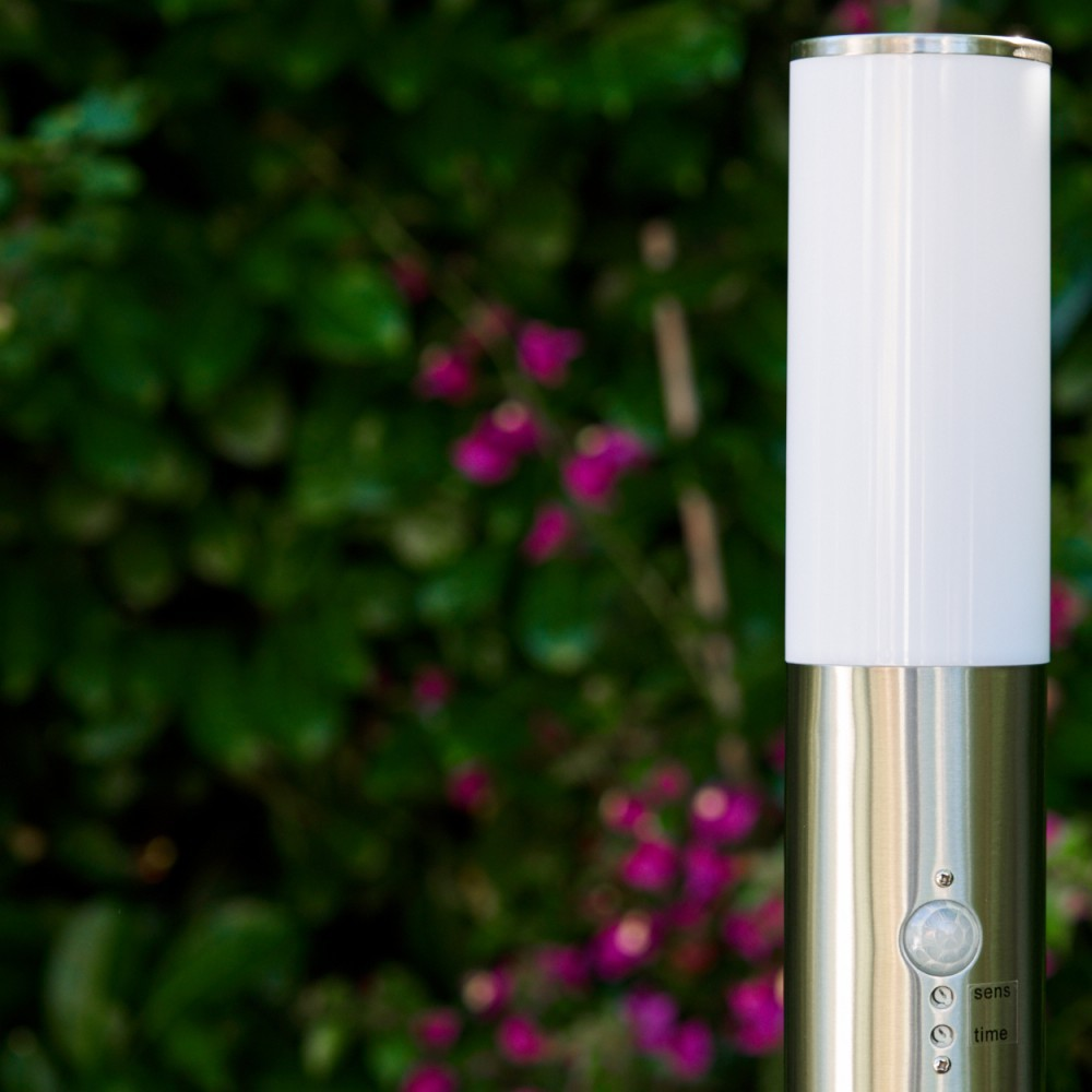 Eine moderne Außenlampe mit weißem Oberteil und silbernem Gehäuse, vor dem Hintergrund grüner Blätter und rosa Blüten.