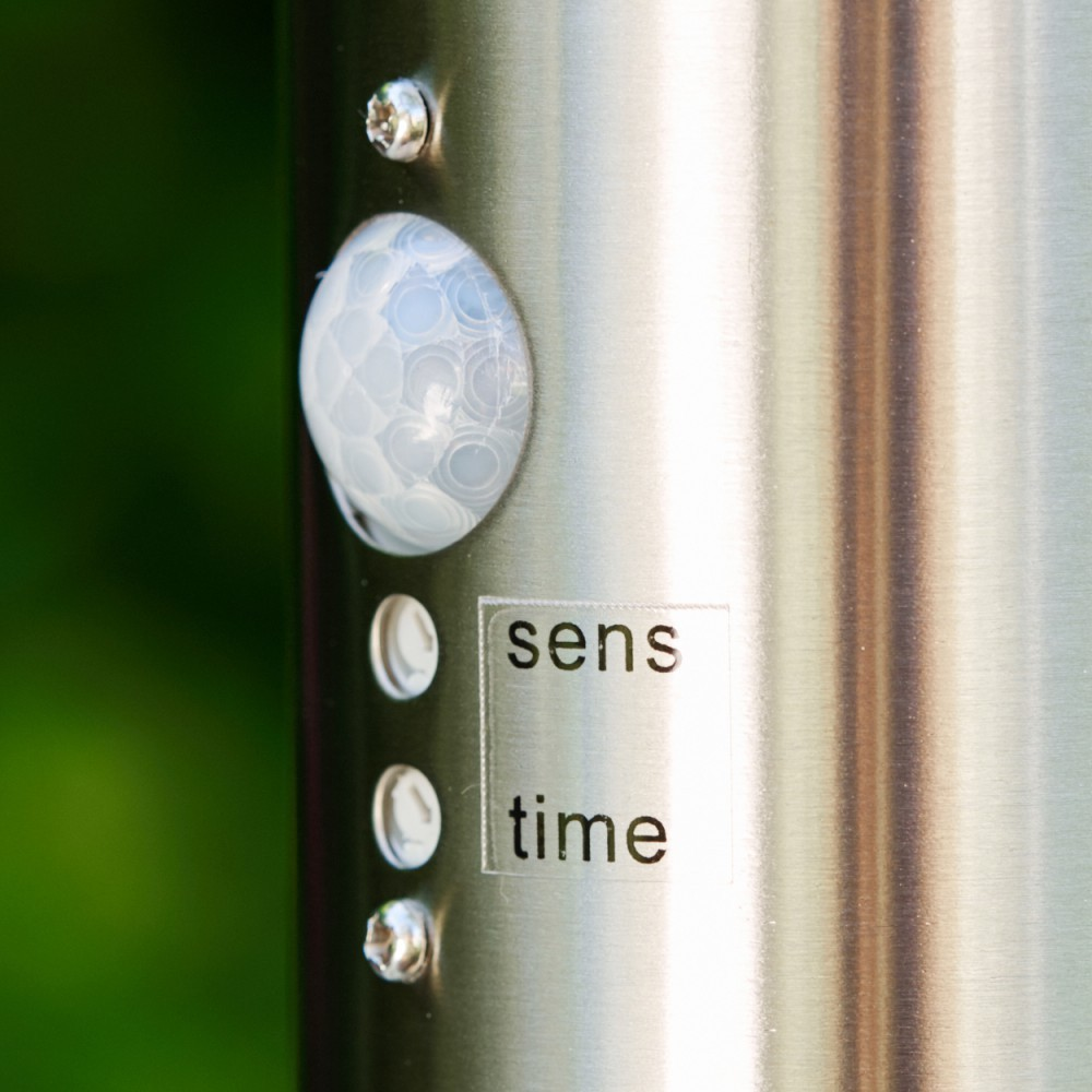 Nahaufnahme einer Metallsäule mit Bewegungsmelder, Knöpfen und Text 'sens time'. Grüner Hintergrund.