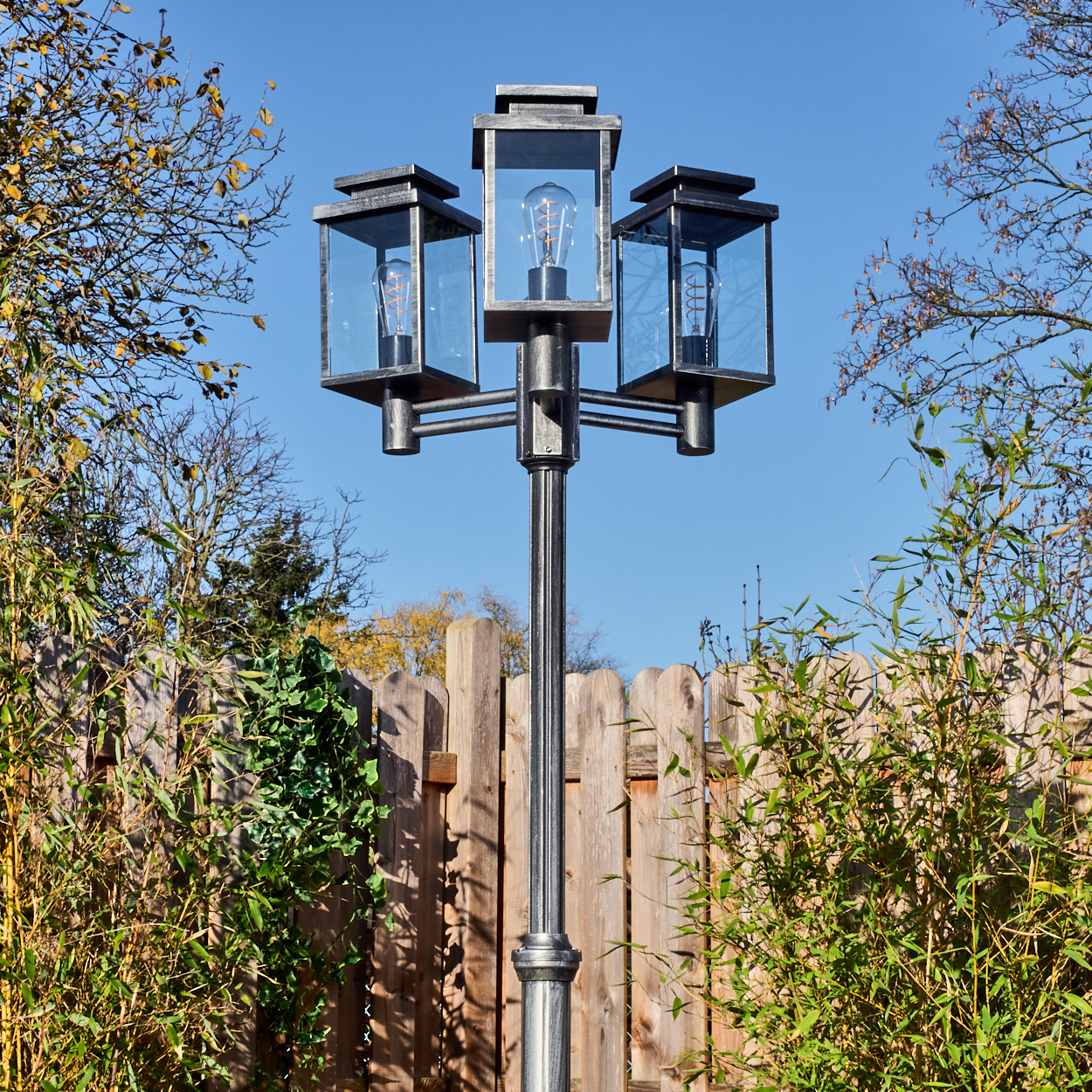 Eine Laterne mit drei Lampen in Glaskästen, vor einem Holzzaun und blauem Himmel. Das Laub umgibt die Laterne.