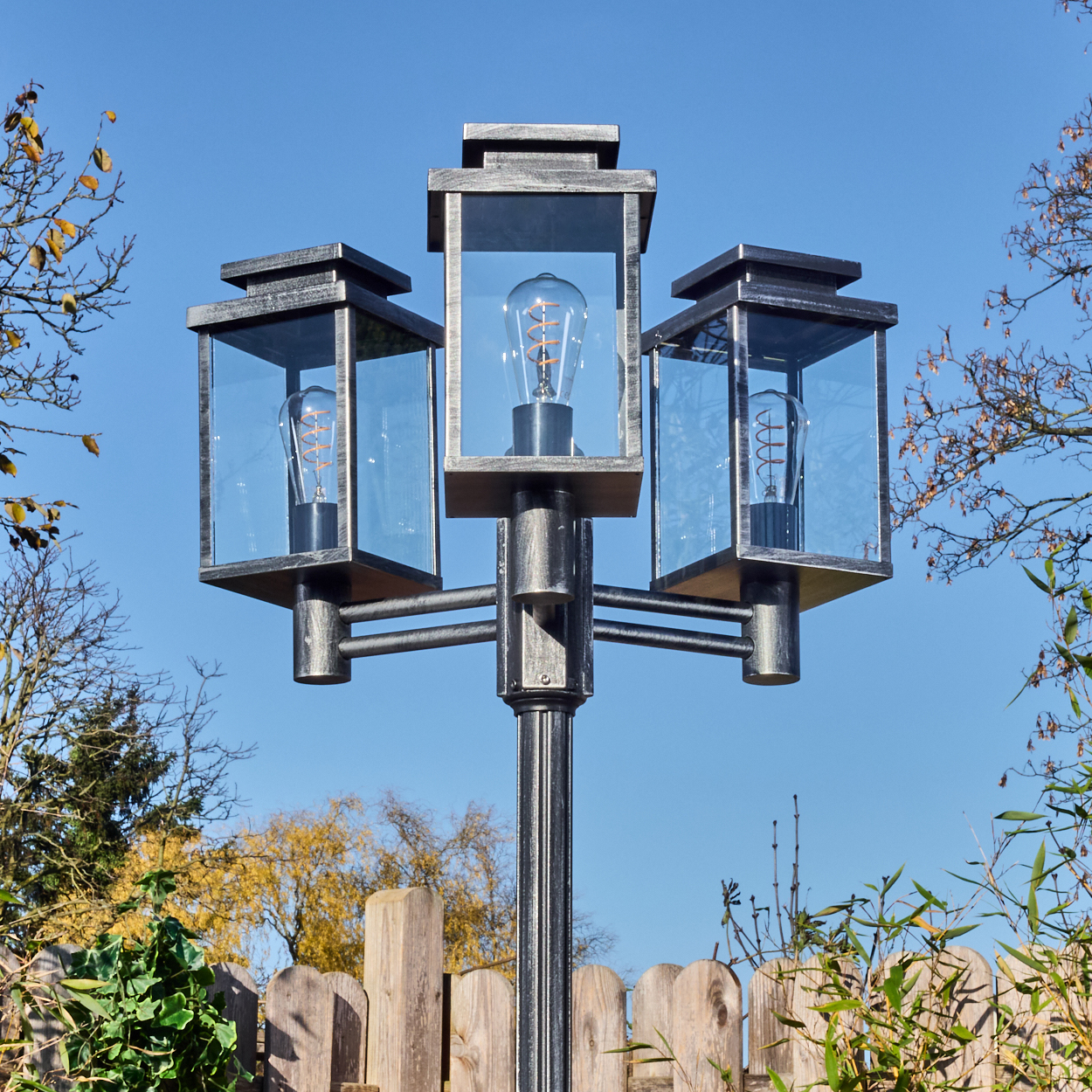 Eine dekorative Außenlampe mit drei Glaseingeschlossenen Lichtern. Sie steht in einem Garten mit Holzzaun und blauem Himmel.