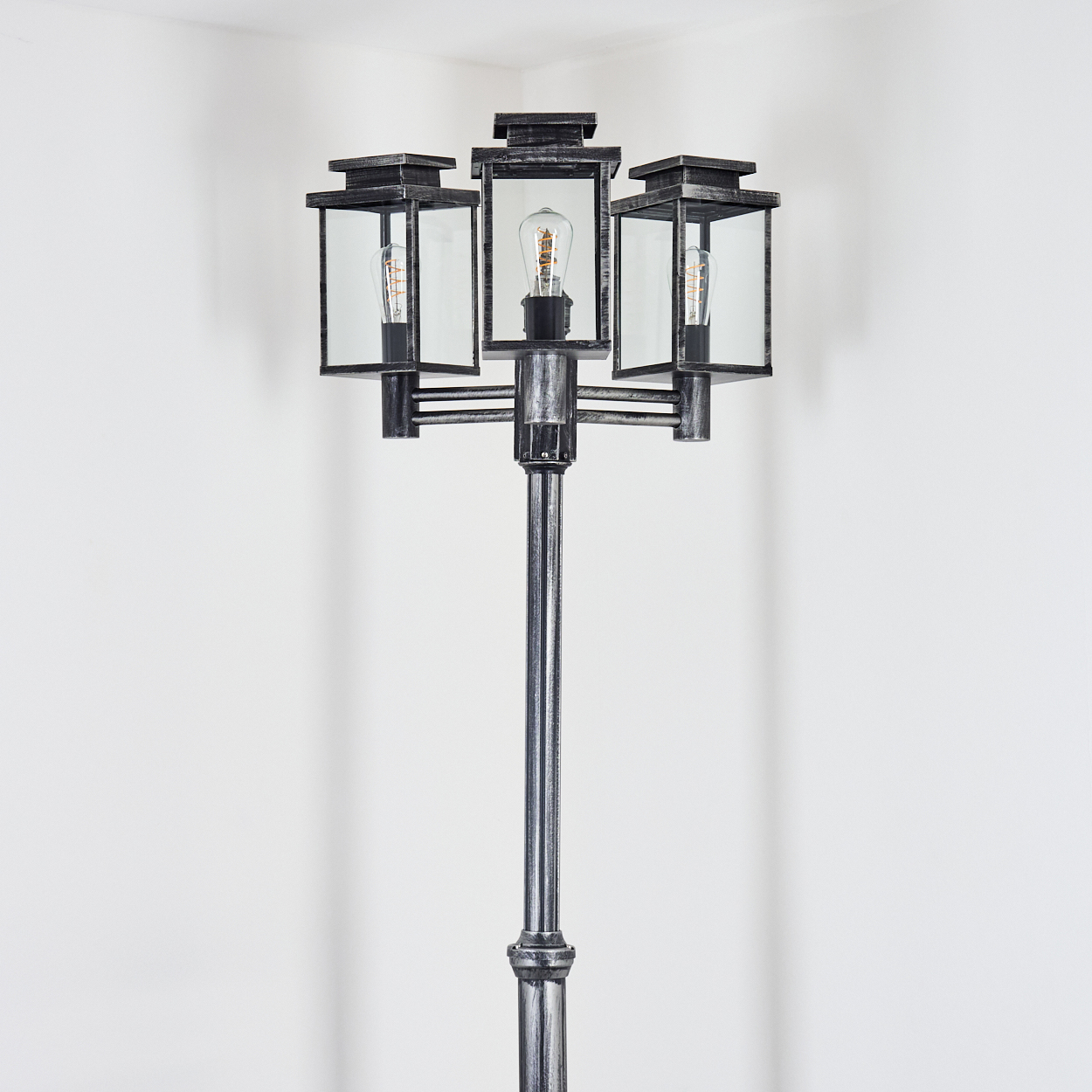 Eine dekorative Außenlampe mit drei Lichtern. Sie hat einen schwarzen Metallrahmen und steht vor einer weißen Wand.
