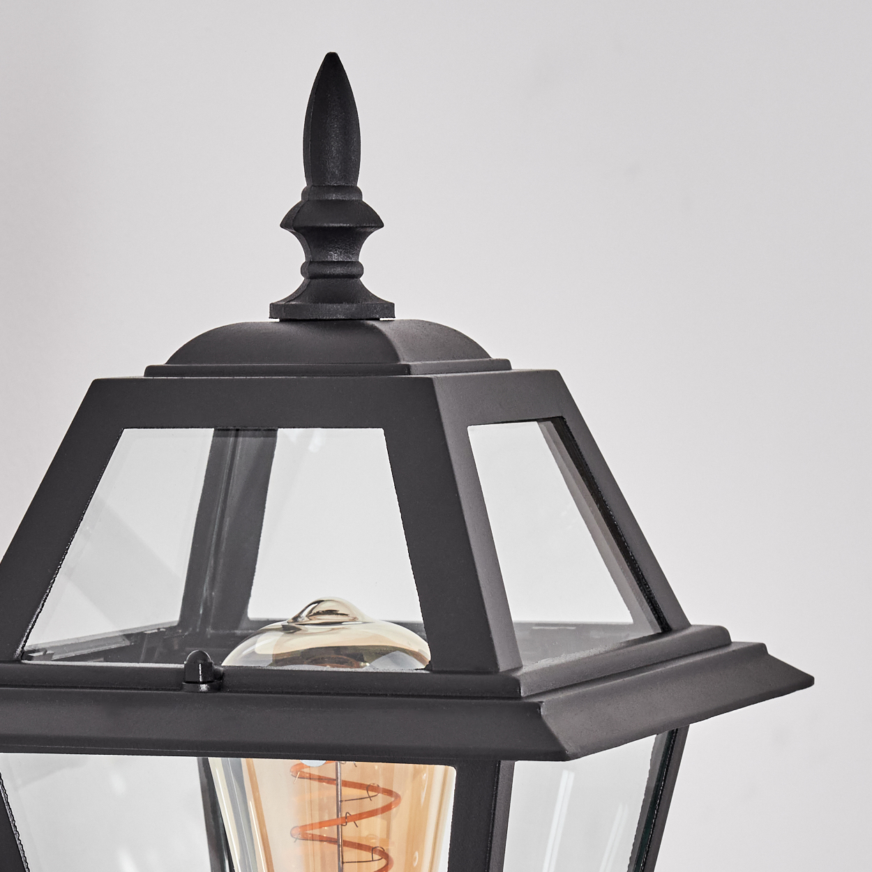 Nahaufnahme einer schwarzen Außenlampe mit Glasscheiben und einem dekorativen Aufsatz. Im Inneren ist eine Glühbirne zu sehen.