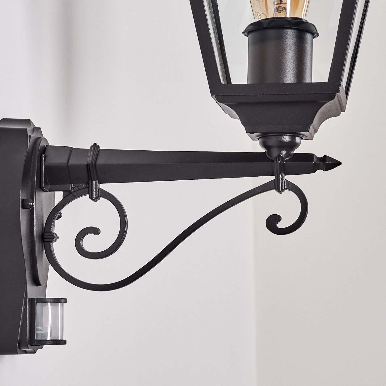 Schwarze Außenlampe. Sie hat ein Glasgehäuse und einen dekorativen Arm mit einem Scroll-Design. Ein Sensor befindet sich an der Wand.