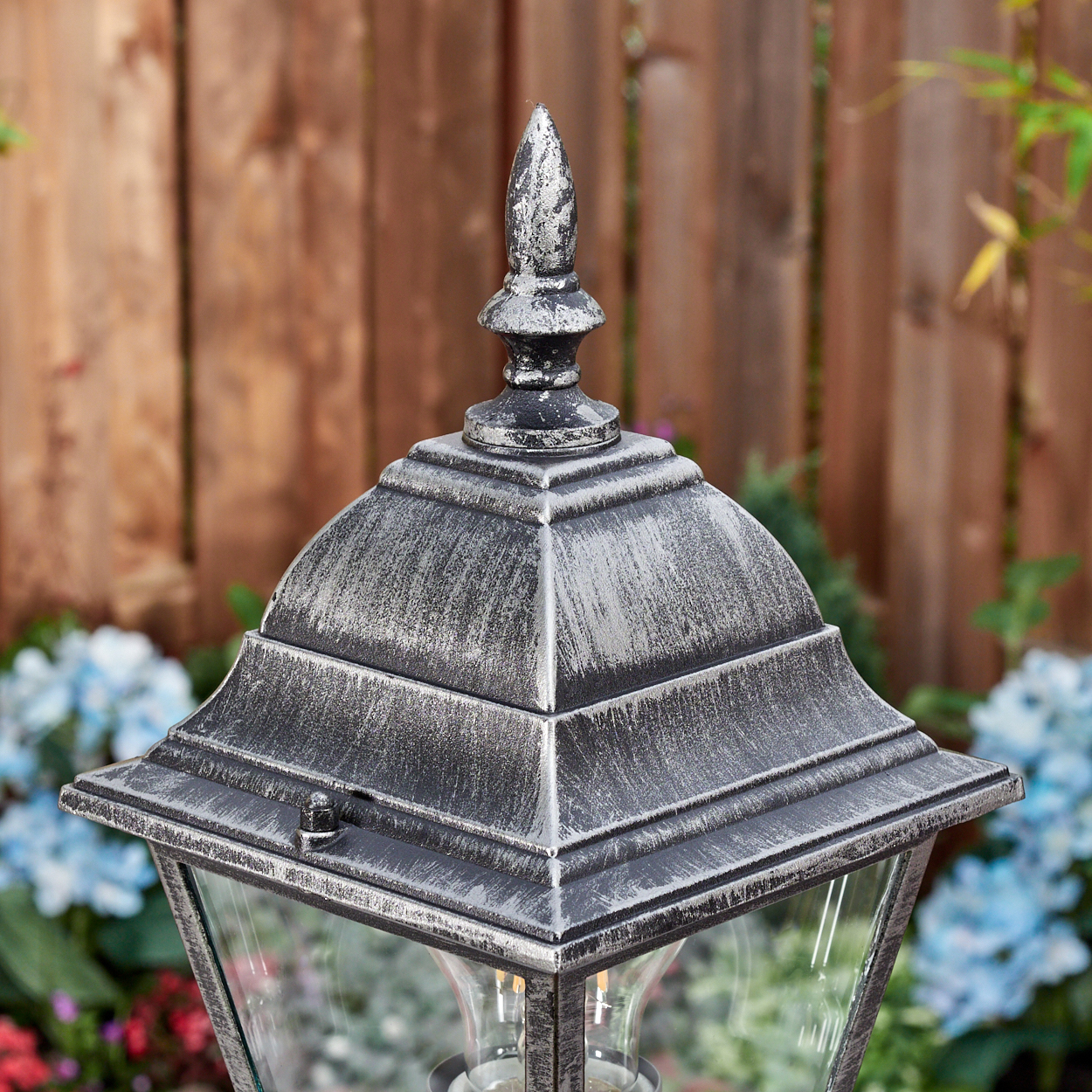 Nahaufnahme einer silbernen Außenlampe mit spitzem Aufsatz. Dahinter ein Holzzaun und blaue Blumen.