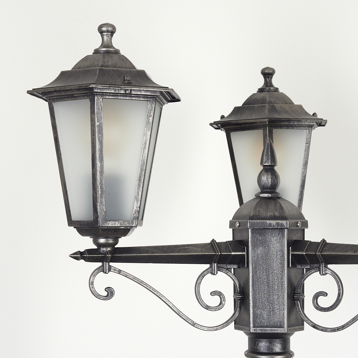 Nahaufnahme einer dekorativen Straßenlaterne. Zwei silberfarbene Laternen mit mattierten Glasschirmen sind auf einem Pfosten vor weißem Hintergrund montiert.