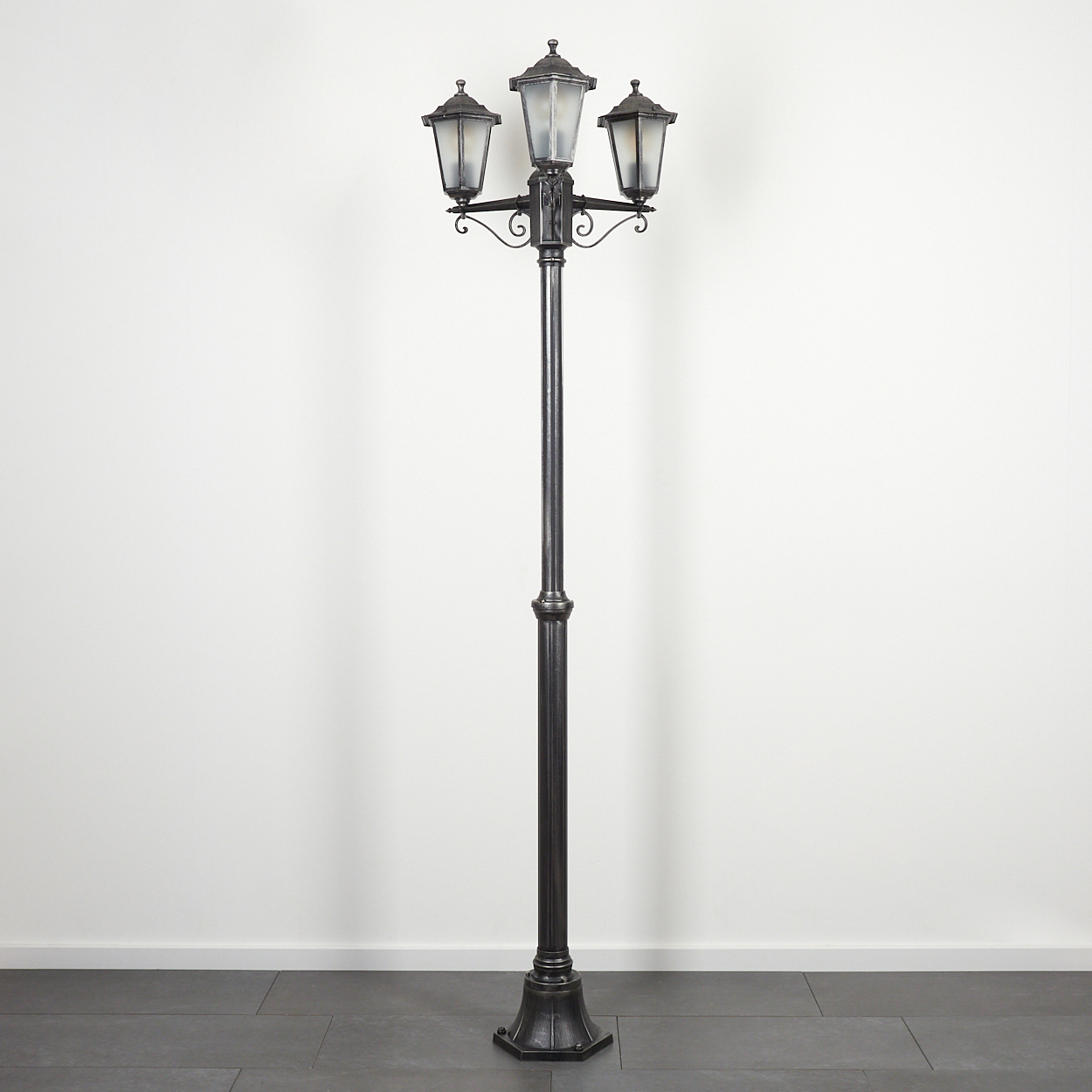 Eine schwarze, verzierte Straßenlaterne mit drei Lichtern steht vor einer weißen Wand und dunklen Fliesen.