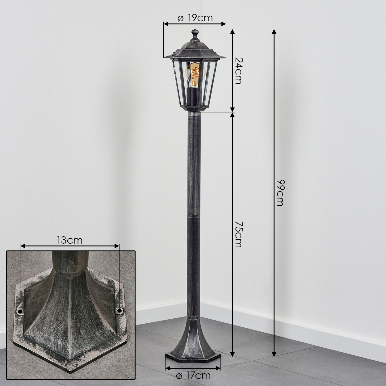 Eine dekorative schwarze und silberne Außenlaterne. Maße werden angezeigt. Die Lampe steht auf einem gefliesten Boden vor einer weißen Wand.