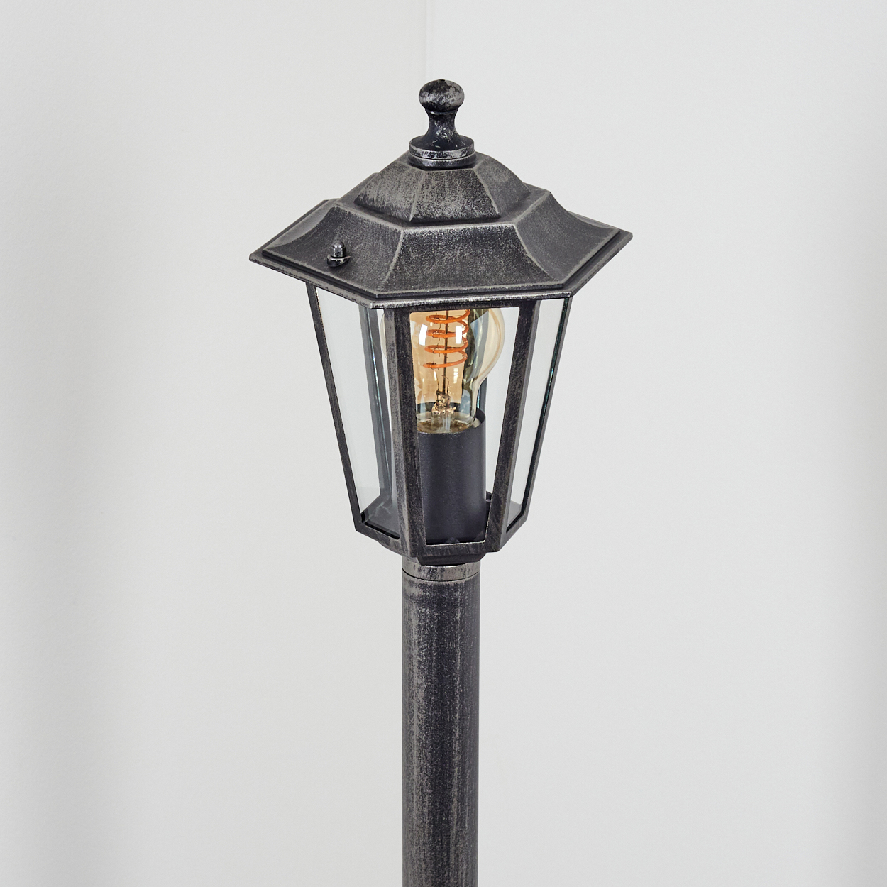 Graue Außenlampe mit Glasgehäuse und sichtbarer Glühbirne, montiert auf einem Pfahl, vor weißem Hintergrund.