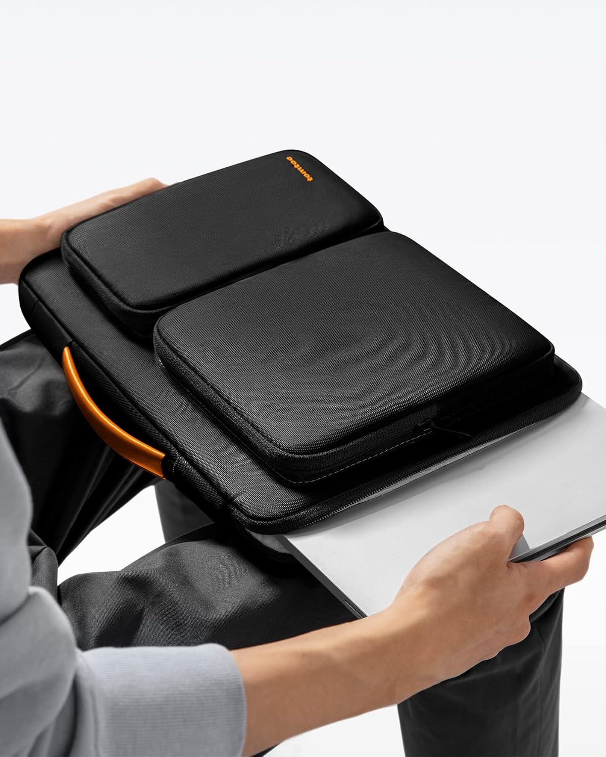 Eine Person hält eine schwarze Laptoptasche mit orangefarbenem Griff. Die Tasche hat drei Fächer und einen Laptop.