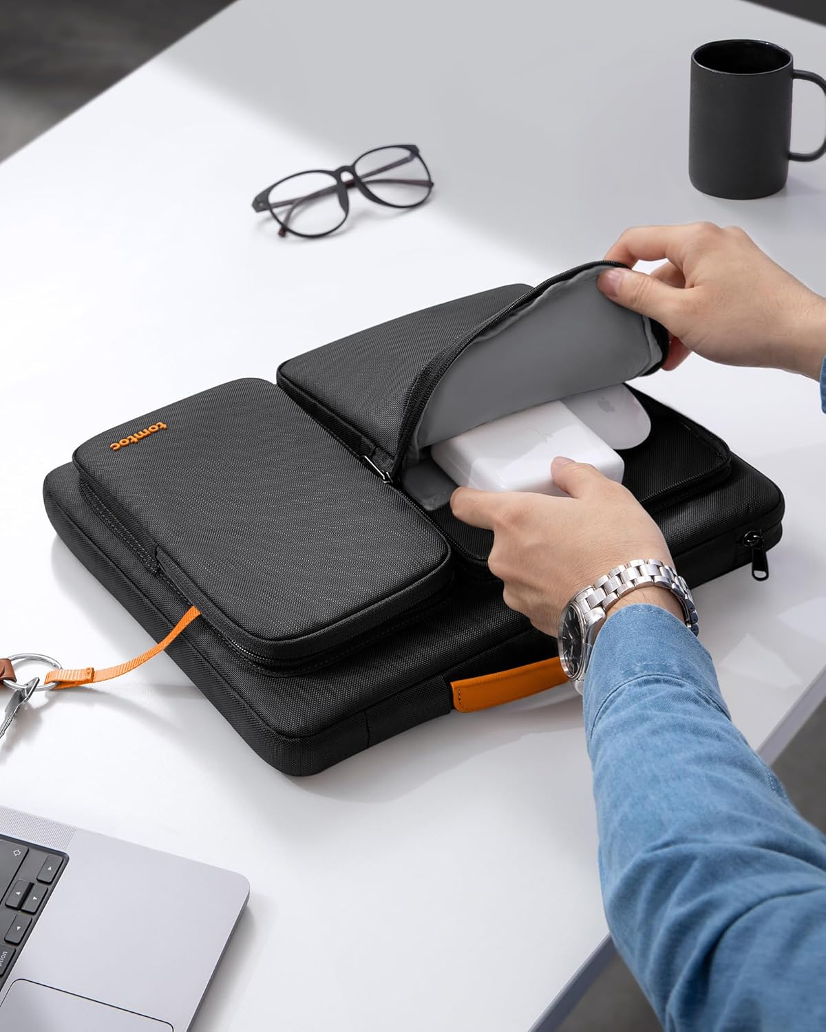 Eine Person öffnet eine schwarze Tasche auf einem weißen Tisch. Darin eine weiße Box. Brille und Becher liegen neben der Tasche.