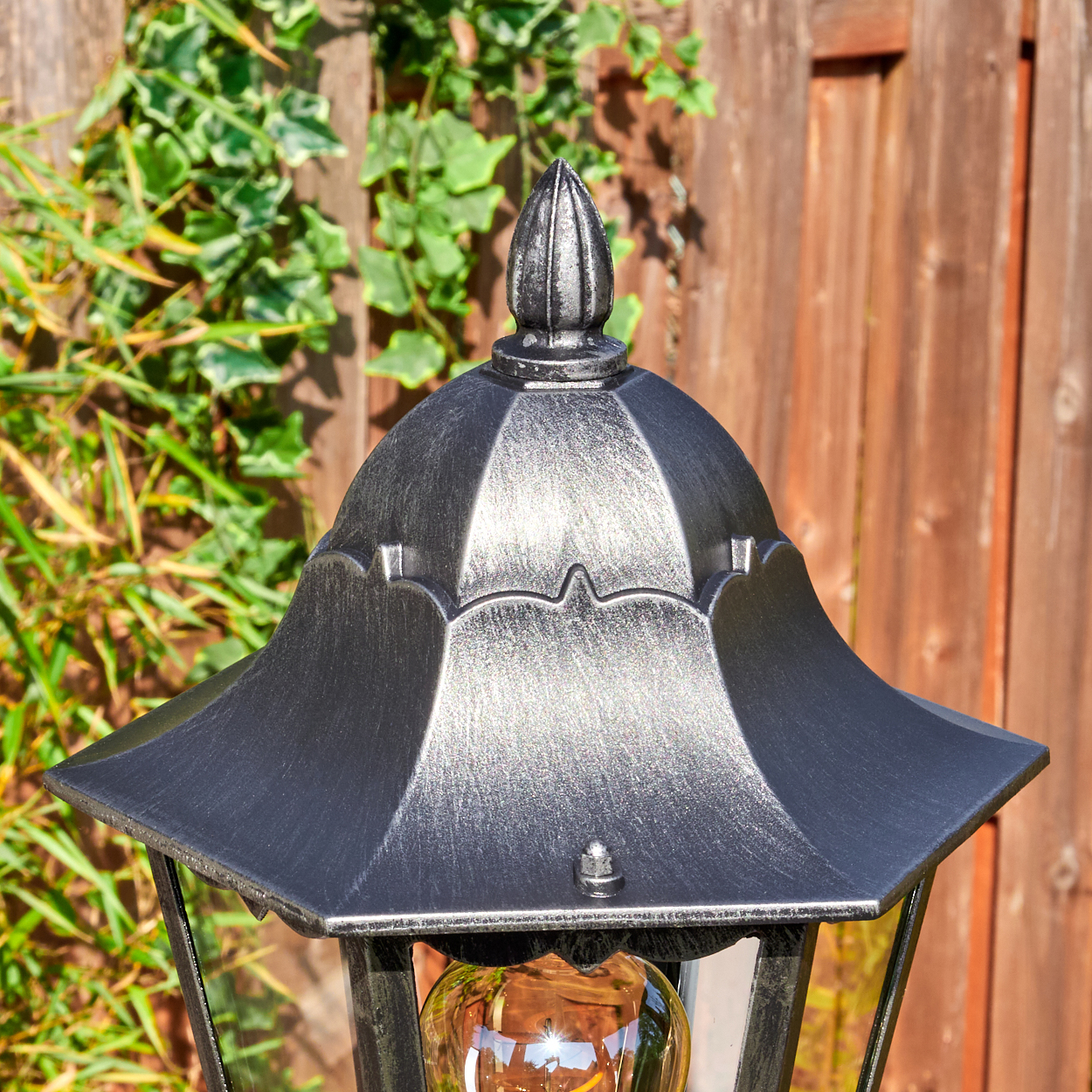 Nahaufnahme einer schwarzen Metall-Außenlampe mit Glühbirne. Efeu und Holzzaun im Hintergrund.