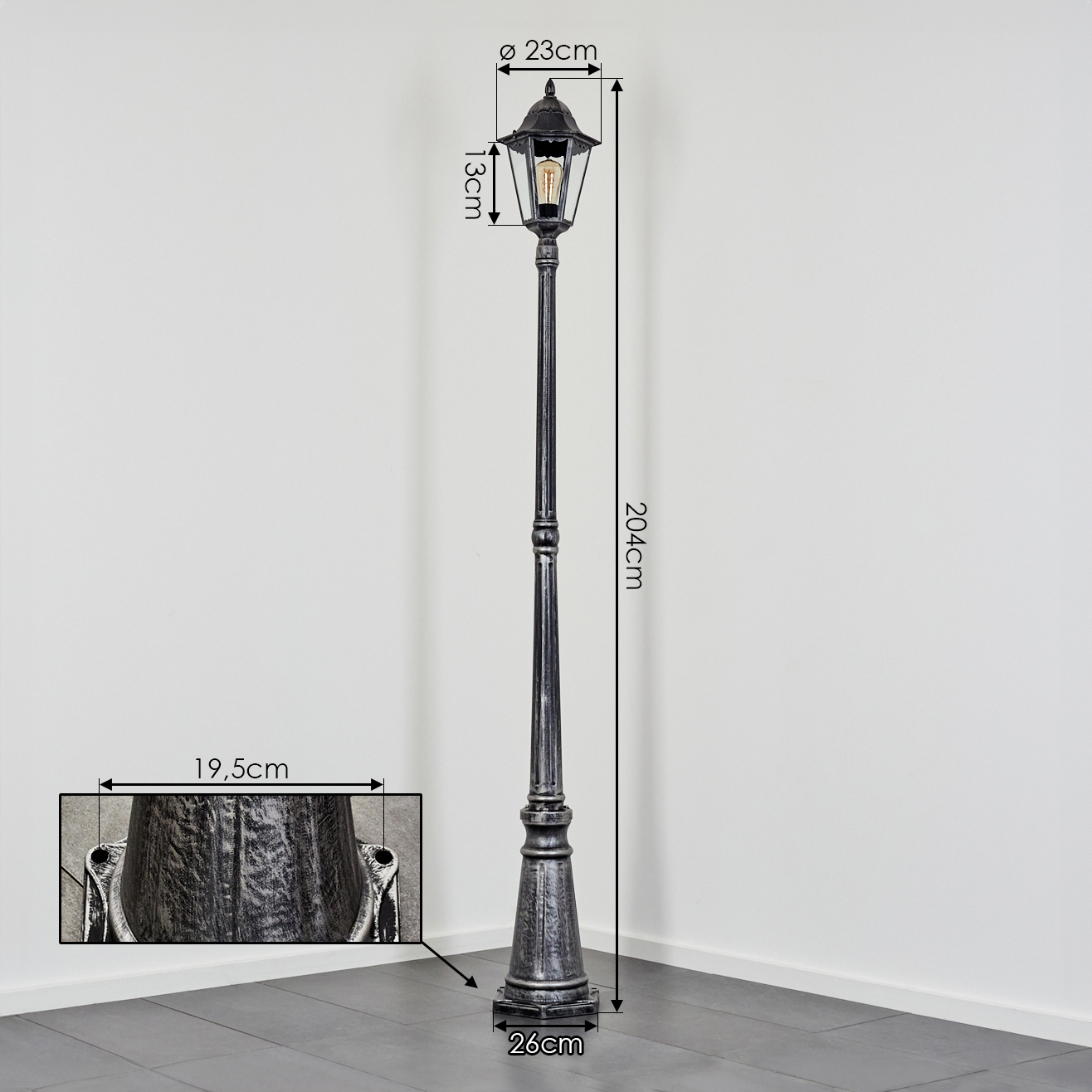 Eine dekorative, schwarze Straßenlaterne mit Maßen. Sie steht in einer Ecke vor einer weißen Wand und einem grauen Boden.