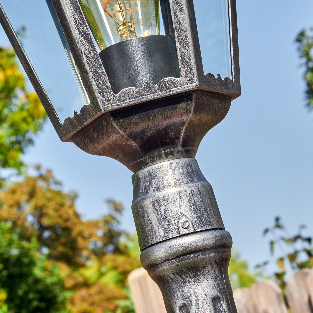 Nahaufnahme einer dekorativen Außenlaterne. Die Lampe hat ein Glasgehäuse und eine Glühbirne im Vintage-Stil. Der Hintergrund ist ein Garten.