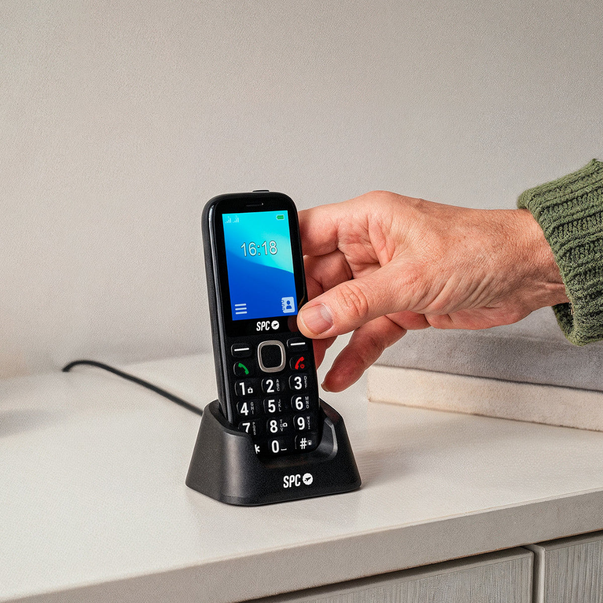 Ein schwarzes Mobiltelefon in seiner Ladestation. Eine Hand hält das Telefon. Der Bildschirm zeigt die Uhrzeit. Der Hintergrund ist ein weißer Tisch.