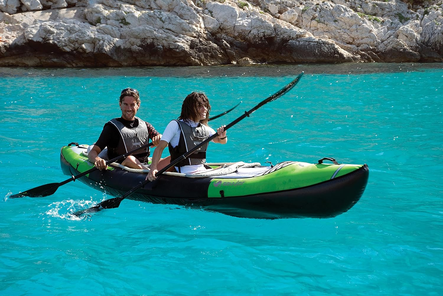 Zwei Personen paddeln in einem grün-schwarzen Kajak auf türkisfarbenem Wasser, mit einer felsigen Küste im Hintergrund.