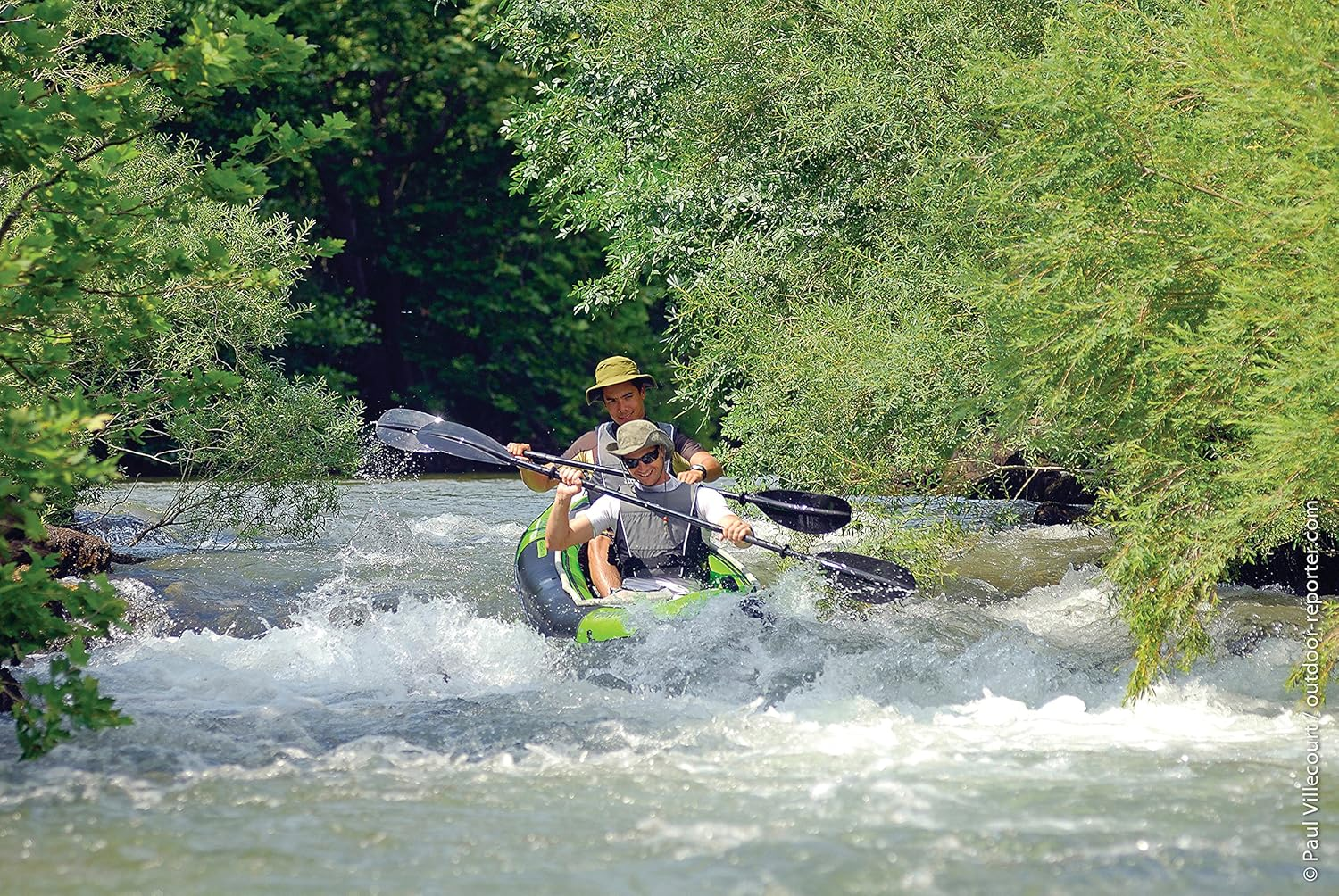 Zwei Männer fahren mit dem Kajak einen Fluss hinunter. Das Kajak ist grün und schwarz. Sie paddeln durch Wildwasser, umgeben von grünen Bäumen.