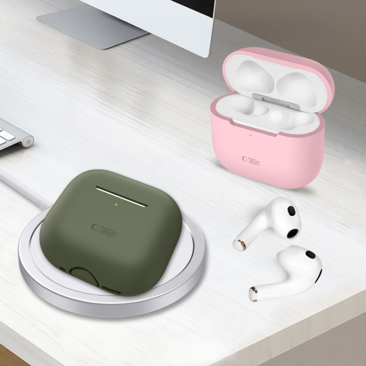 Eine olivgrüne AirPod-Hülle auf einem Ladepad, eine rosa Hülle und weiße AirPods liegen auf einem weißen Schreibtisch.