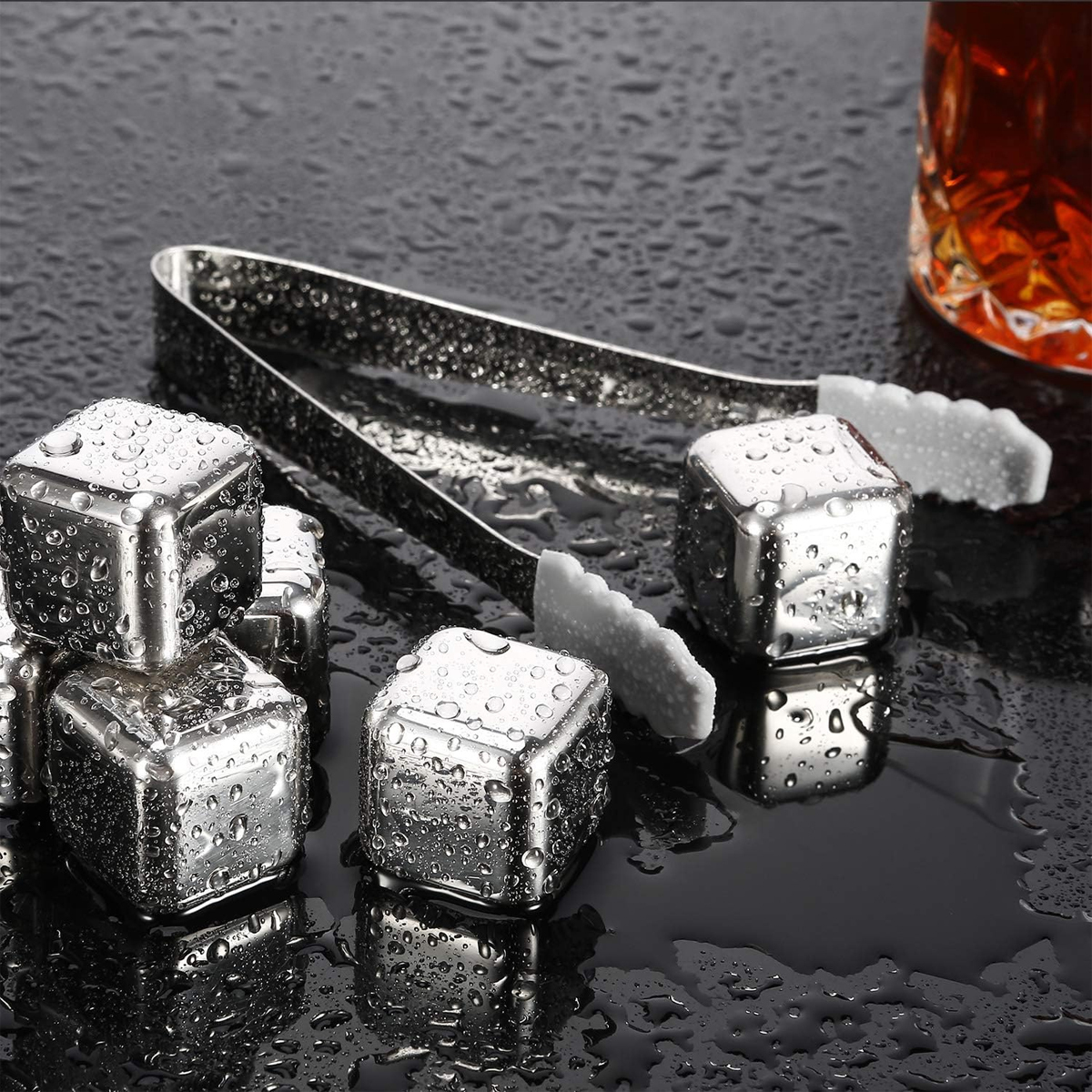 Edelstahl-Eiswürfel mit Zange auf nasser, dunkler Oberfläche. Im Hintergrund ein Glas mit bernsteinfarbener Flüssigkeit.
