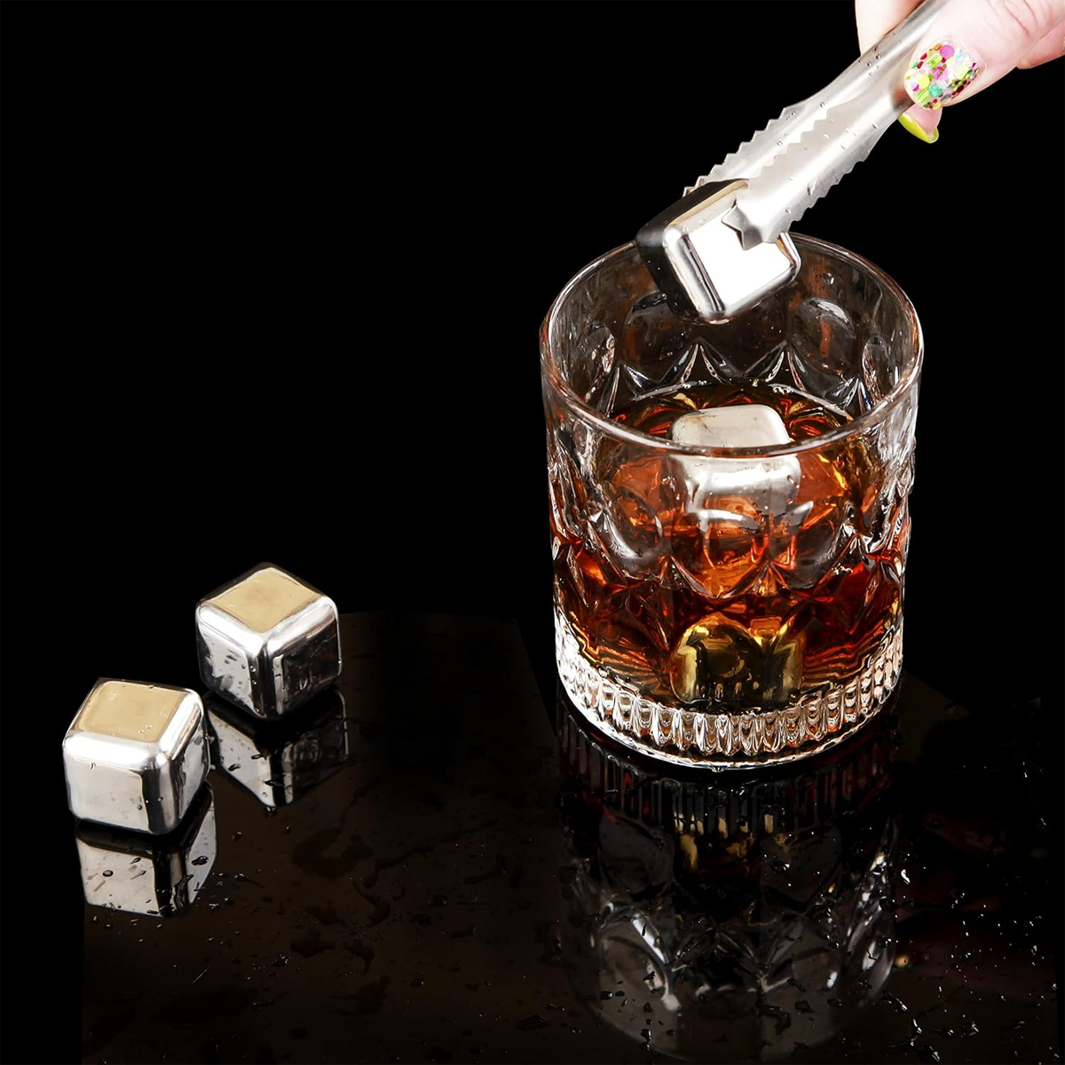 Eine Hand mit einer Zange legt einen Metall-Eiswürfel in ein Glas mit bernsteinfarbener Flüssigkeit. Zwei Würfel liegen auf der glänzenden schwarzen Oberfläche.