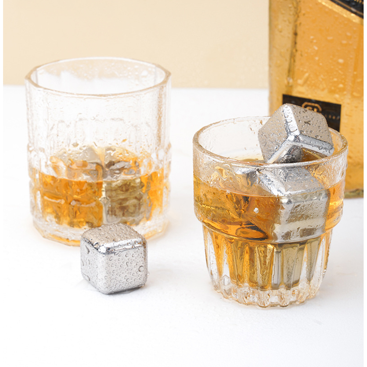 Zwei Gläser mit Whiskey und Metall-Eiswürfeln. Ein Würfel ist separat. Eine Flasche ist im Hintergrund.