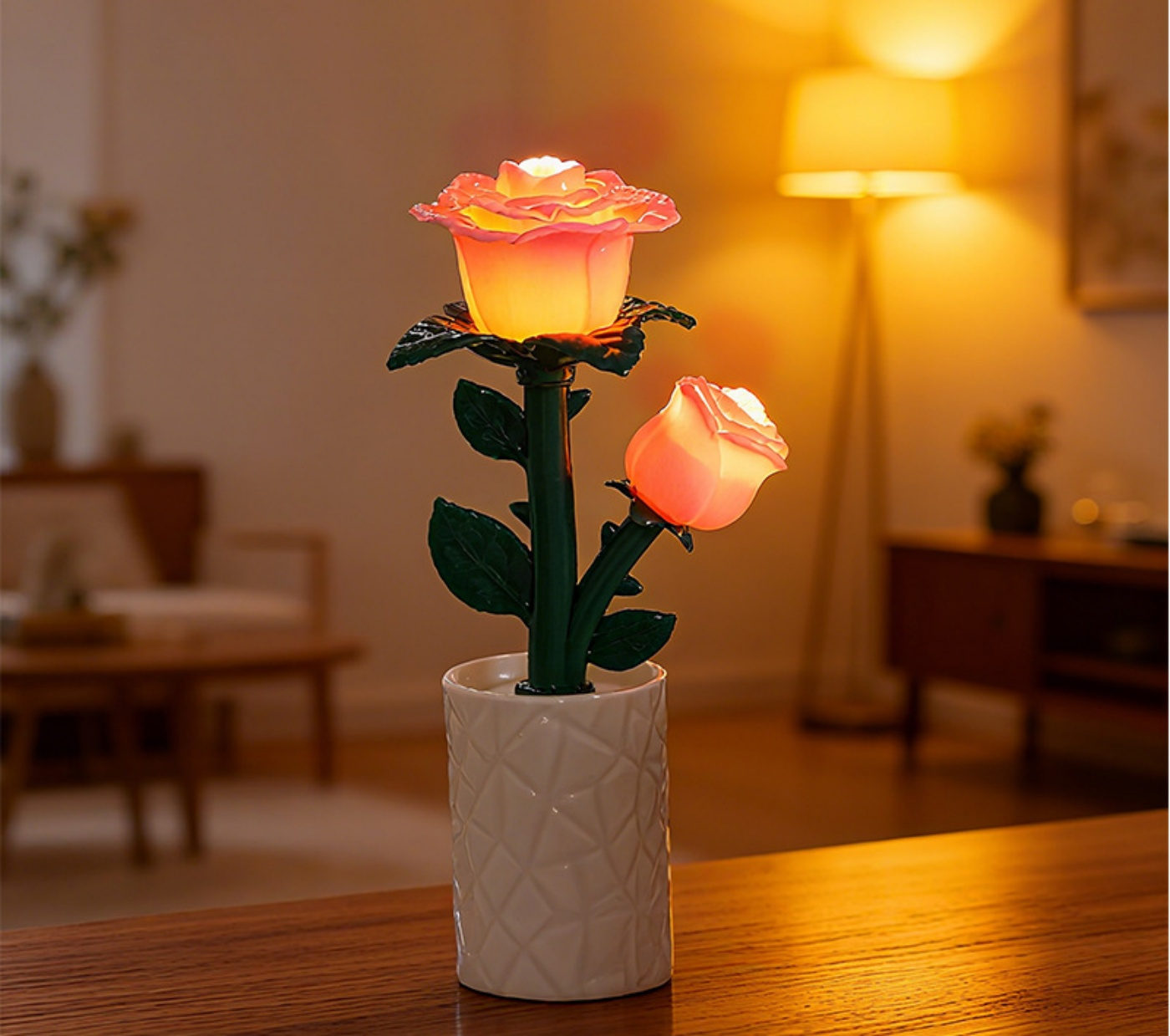 Eine rosenförmige Lampe mit zwei Blüten in einer weißen Vase auf einem braunen Tisch. Im Hintergrund eine Stehlampe und Möbel.