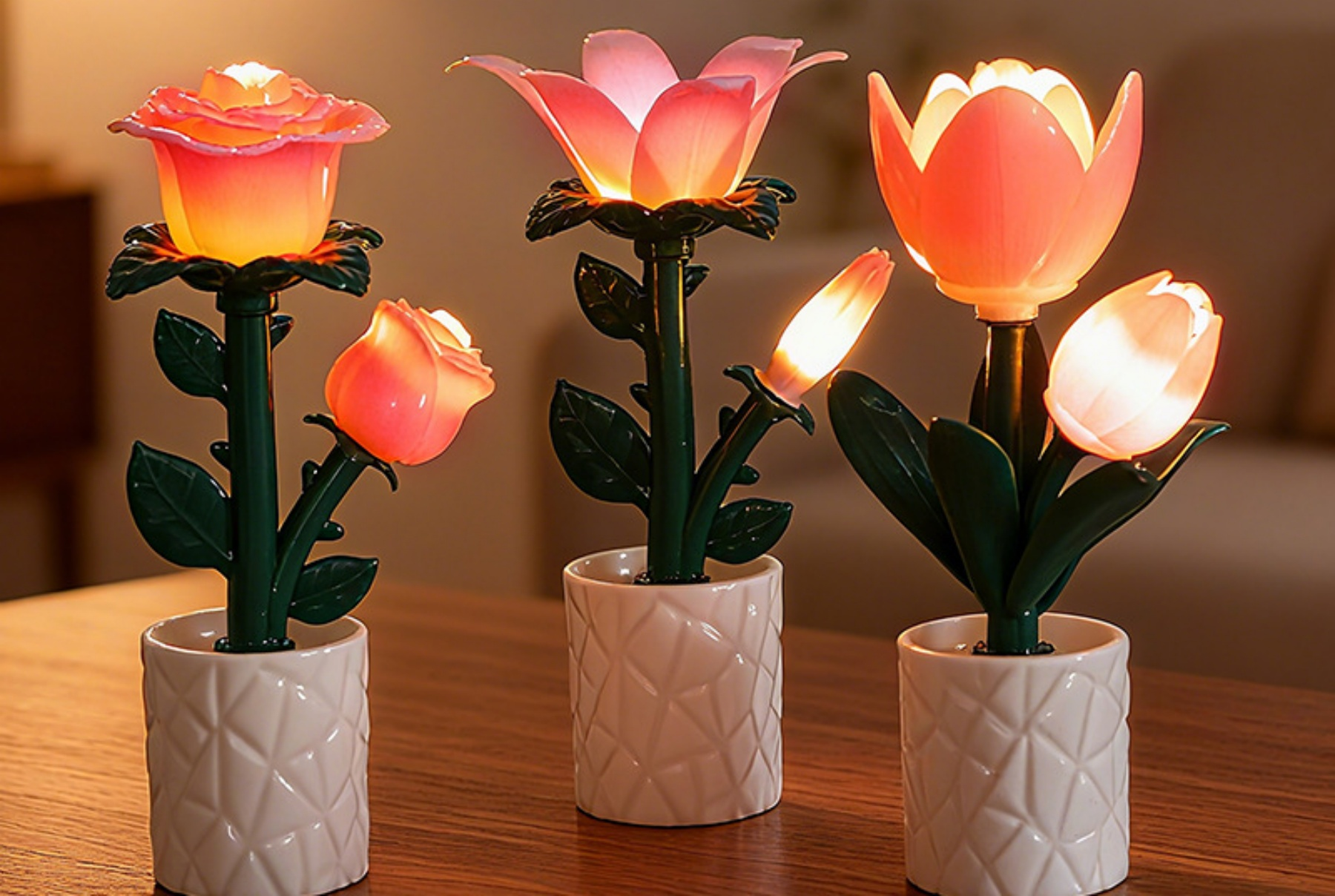 Drei blütenförmige Lampen auf einem Holztisch. Jede hat einen weißen Topf. Die Blumen sind rosa und orange. Grüne Stiele und Blätter.