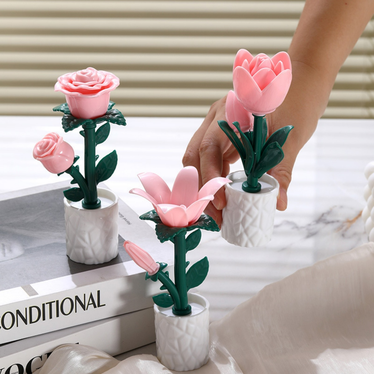 Rosa Kunstblumen in weißen Töpfen. Eine Hand hält eine. Der Hintergrund umfasst Bücher und eine weiße Oberfläche.