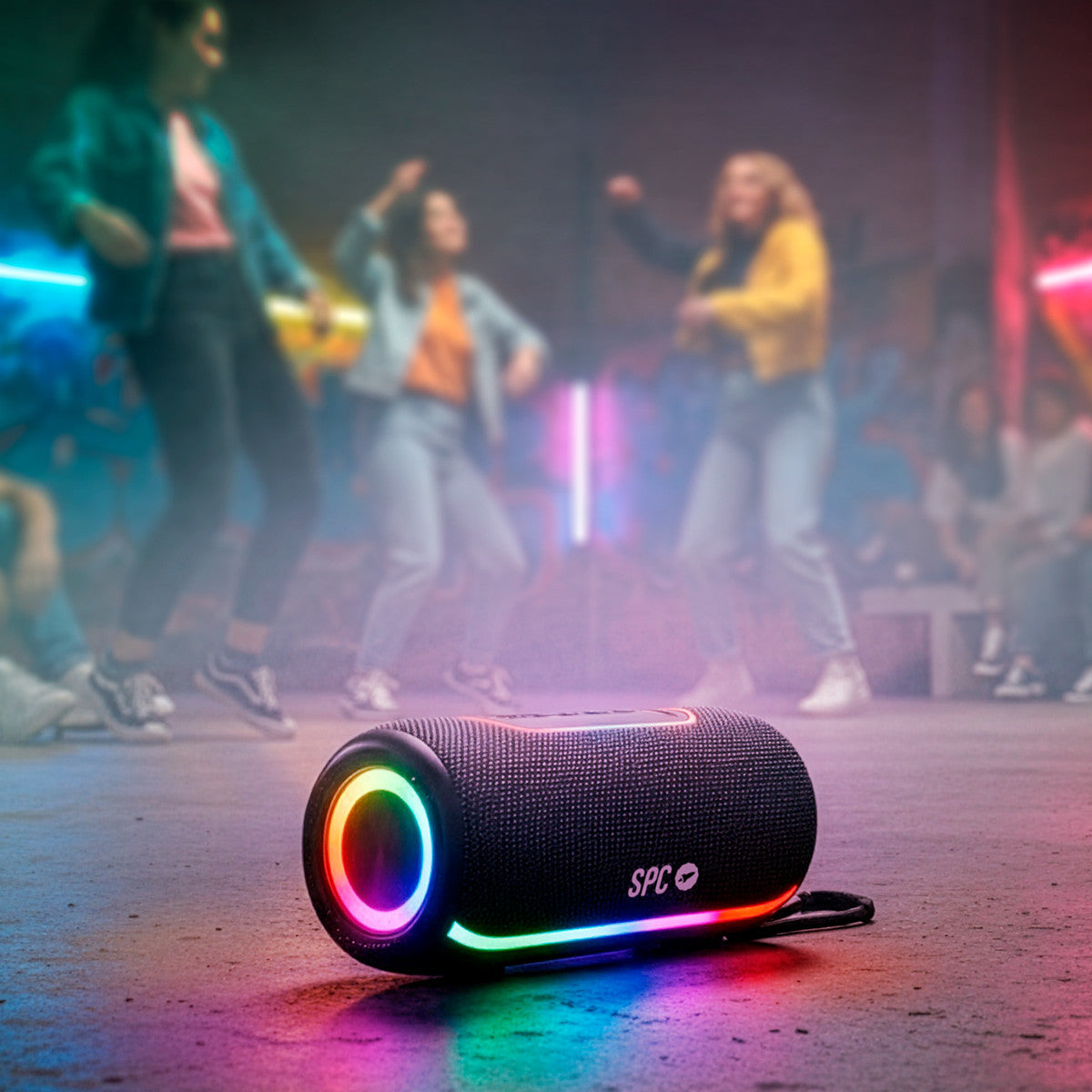 Un altoparlante nero con luci colorate è appoggiato su una superficie. Persone danzanti sullo sfondo. Luci colorate illuminano la scena.