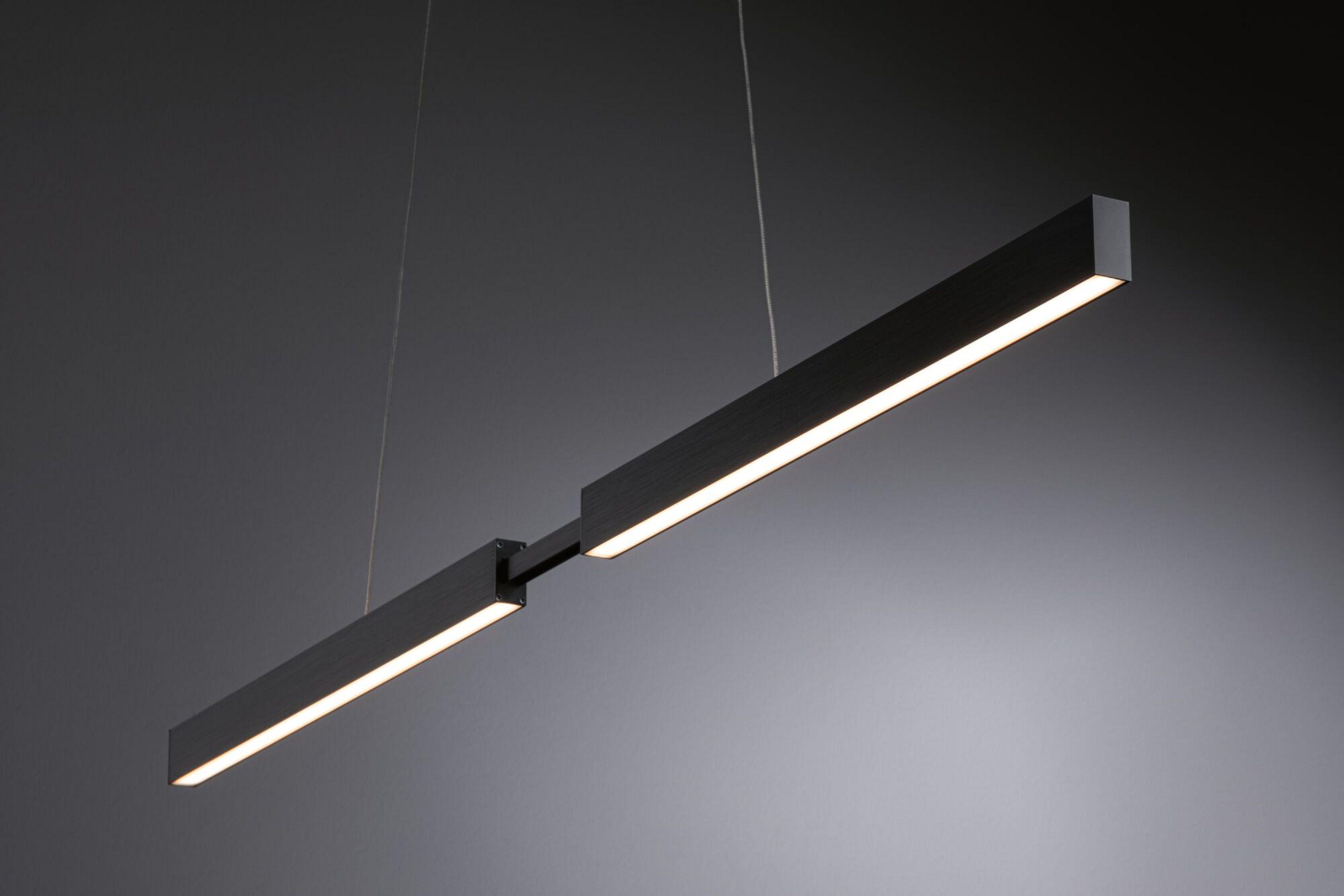 Un luminaire linéaire noir moderne est suspendu au plafond sur un fond gris. Il a une lumière blanche et vive.