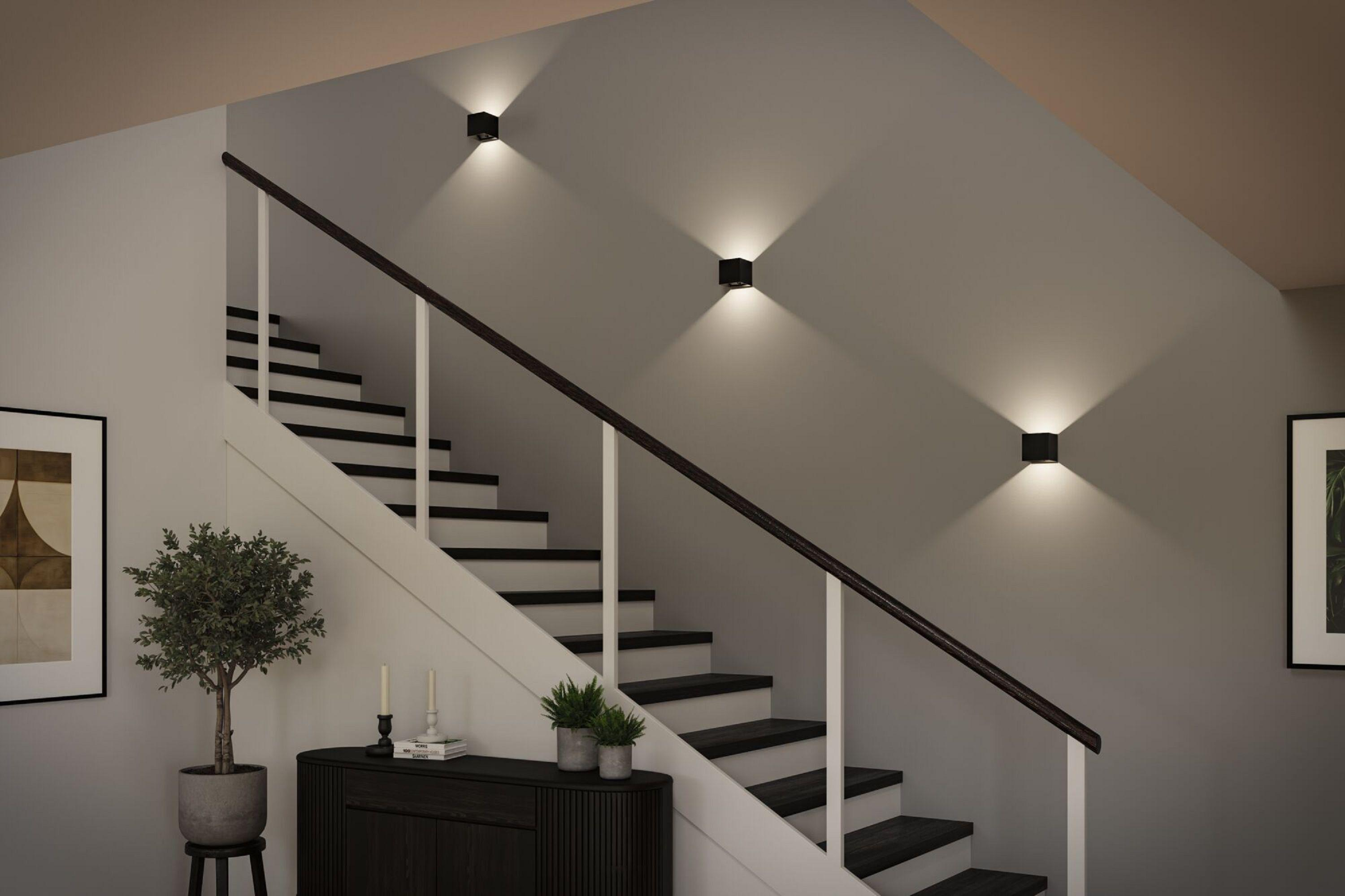 Schody z czarnymi stopniami, białymi podstopniami i drewnianą poręczą. Kinkiety oświetlają przestrzeń.
