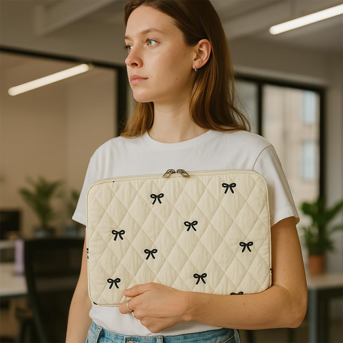 Eine Frau hält eine cremefarbene Laptoptasche mit schwarzen Schleifen. Sie trägt ein weißes Hemd und blickt in einer Büroumgebung nach links.