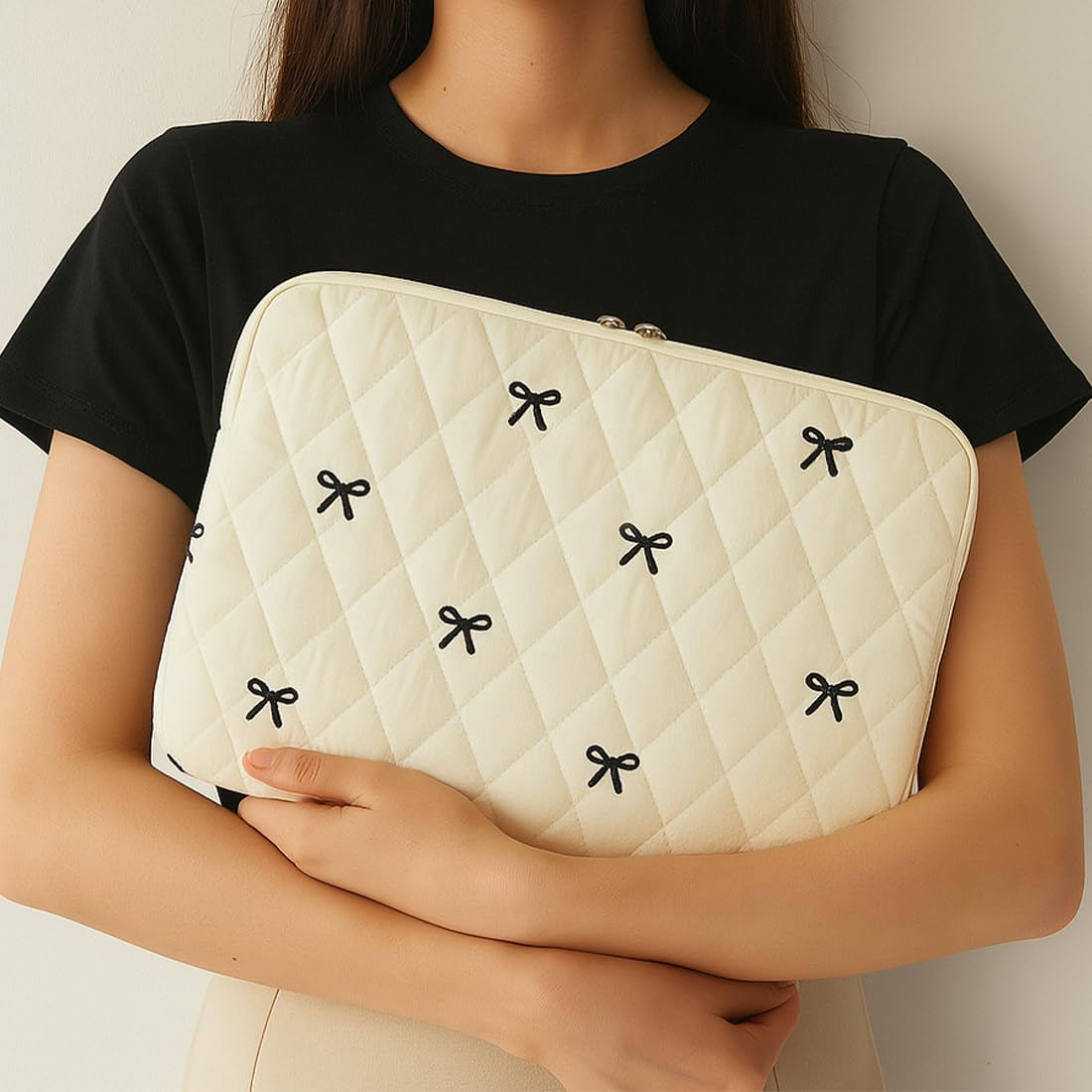 Eine Person hält eine cremefarbene Laptoptasche mit schwarzen Schleifen. Sie trägt ein schwarzes Hemd und beige Hose.