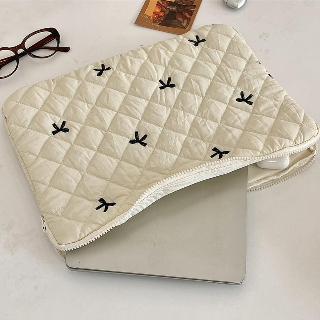 Eine cremefarbene Laptoptasche mit schwarzer Schleifenstickerei, geöffnet, um einen Laptop zu enthüllen. Daneben liegen eine Brille, eine Karte und ein weißes Kopfhörergehäuse.