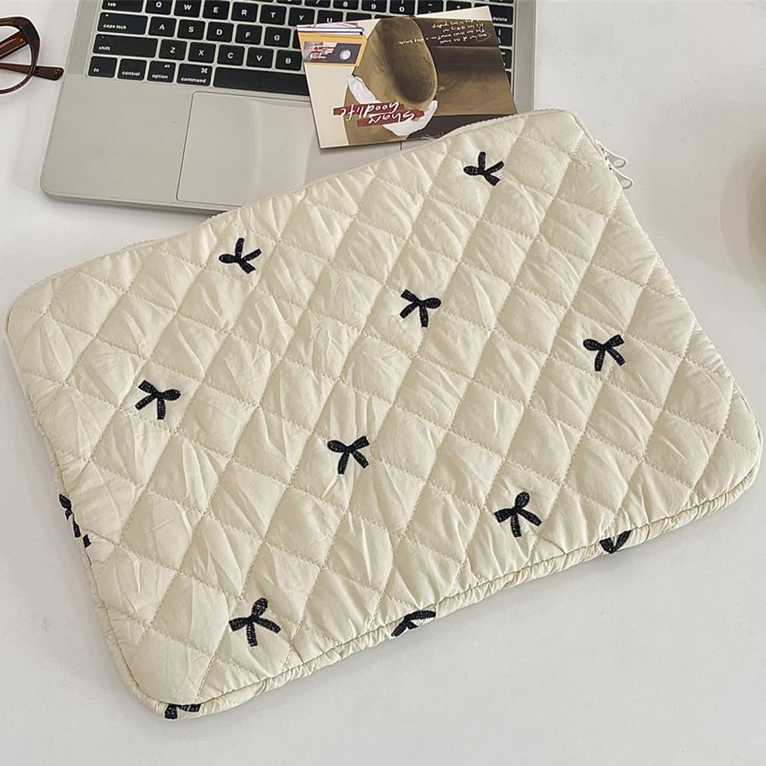 Eine cremefarbene Laptoptasche mit schwarzen Schleifenmustern liegt auf einer weißen Oberfläche. Ein Laptop, eine Brille und eine Karte sind im Hintergrund.
